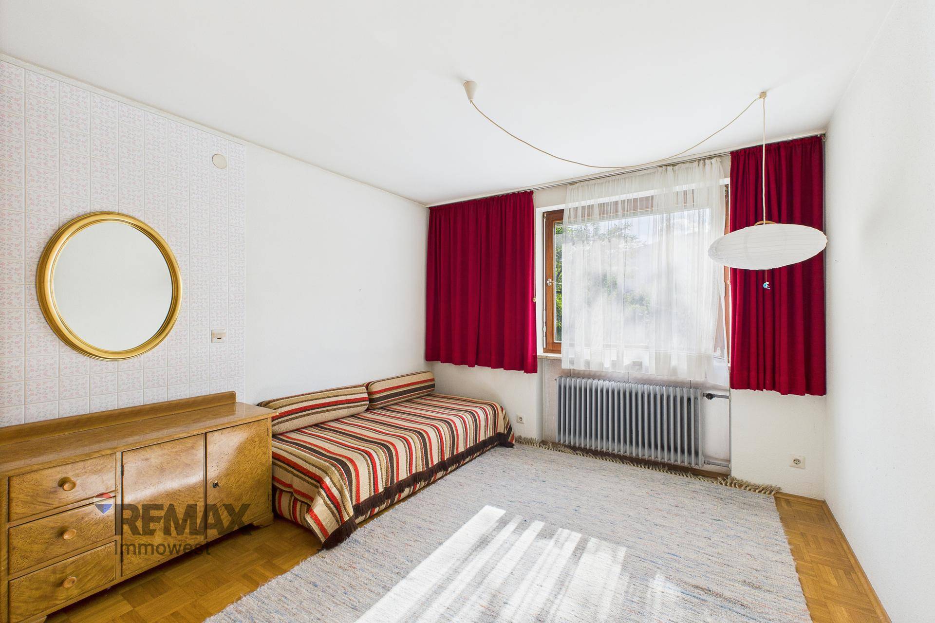 Schlafzimmer mit Einzelbett, Holzmöbeln, rotem Vorhang und hellem Parkettboden.