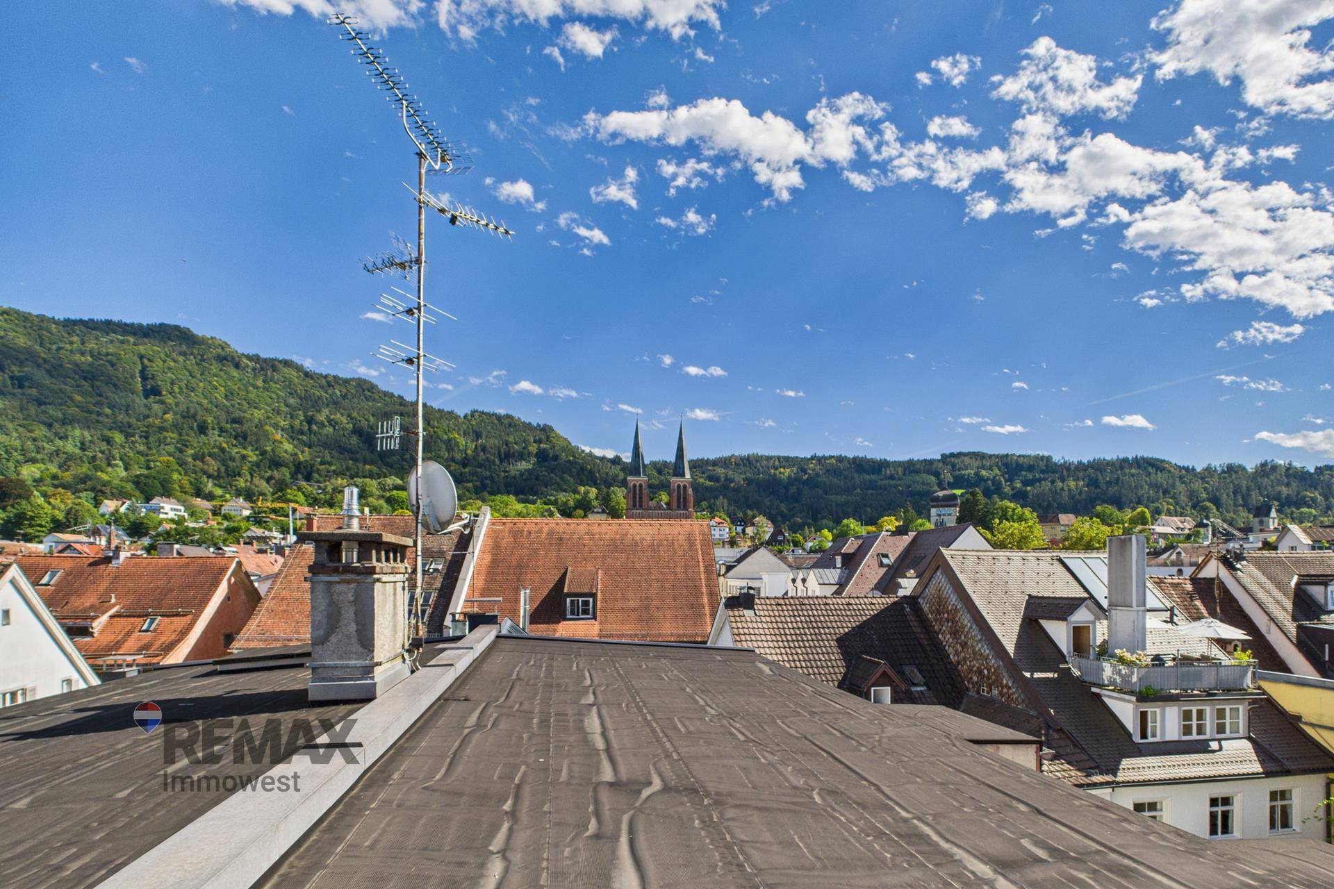 Panoramablick über die Dächer der Stadt mit grünen Hügeln im Hintergrund und blauem Himmel.