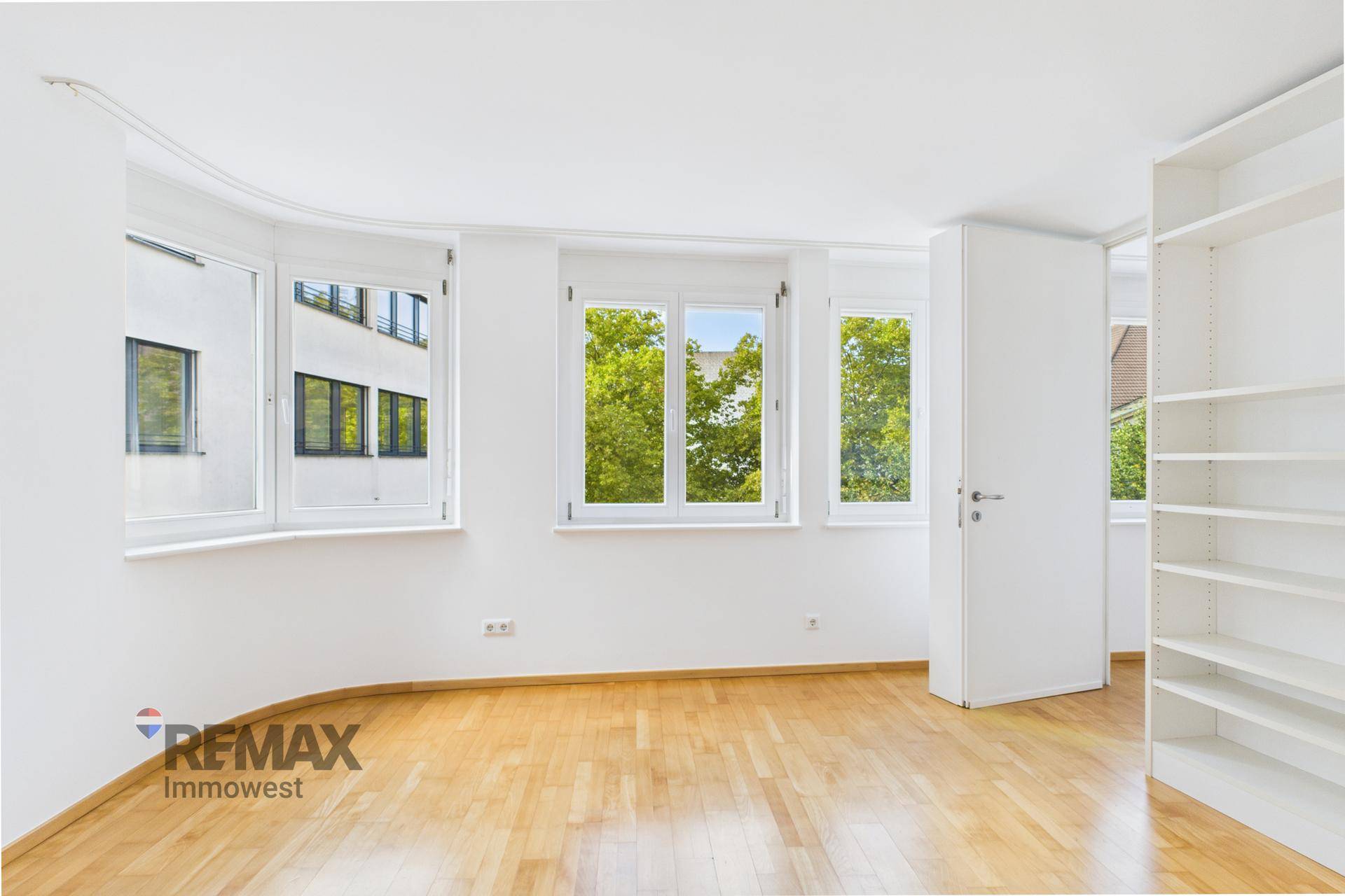 Helles Zimmer mit Holzboden und mehreren Fenstern, die einen Blick ins Grüne ermöglichen.