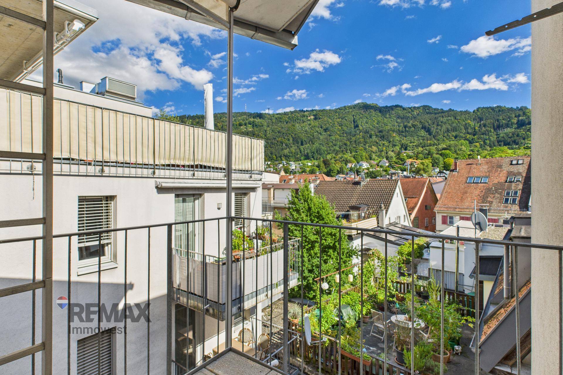 Blick vom Balkon auf einen begrünten Innenhof und die umliegende Berglandschaft unter blauem Himmel.