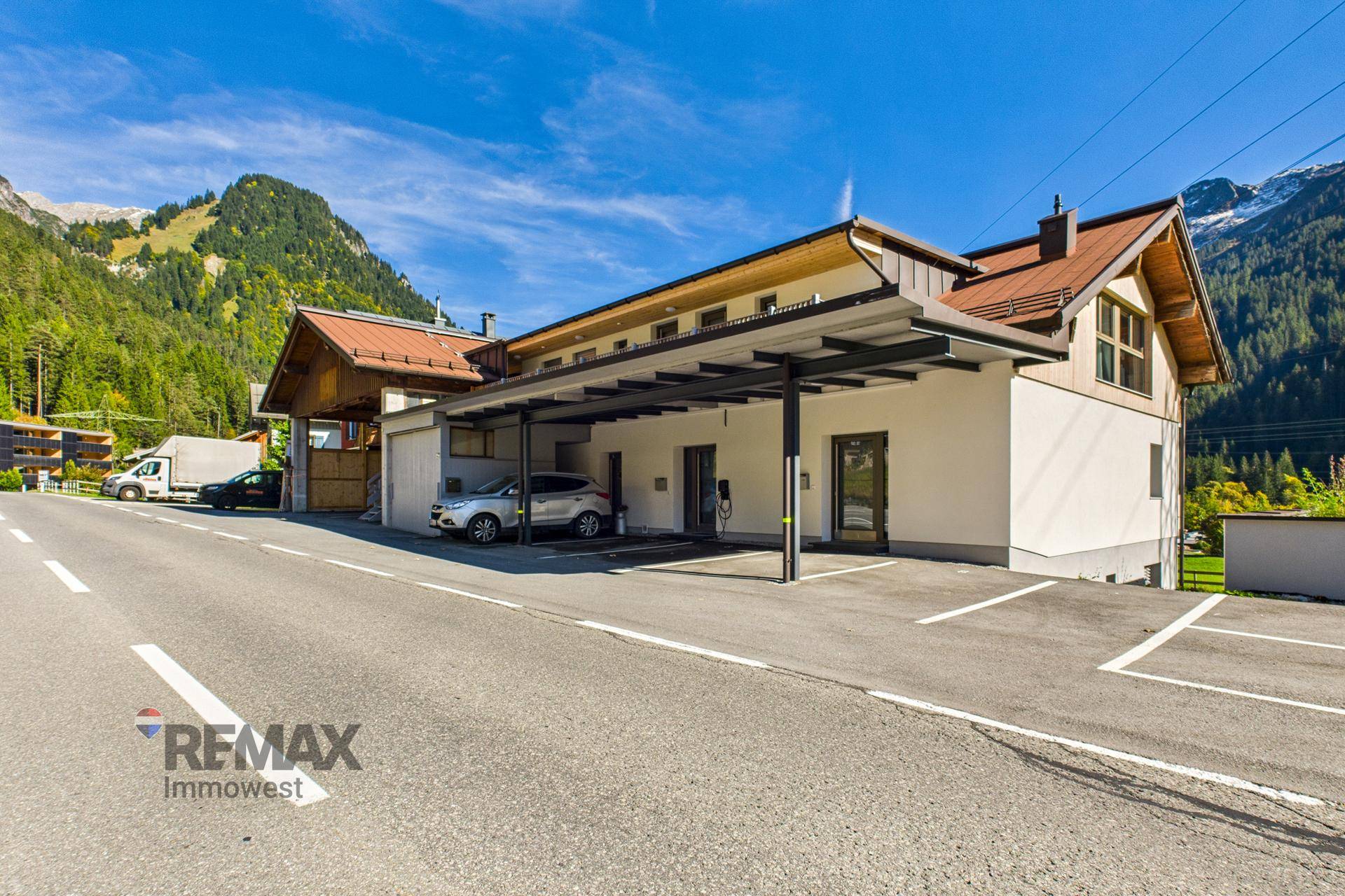 Modernes Gebäude mit Carport und Parkplätzen, umgeben von grünen Bergen unter blauem Himmel.