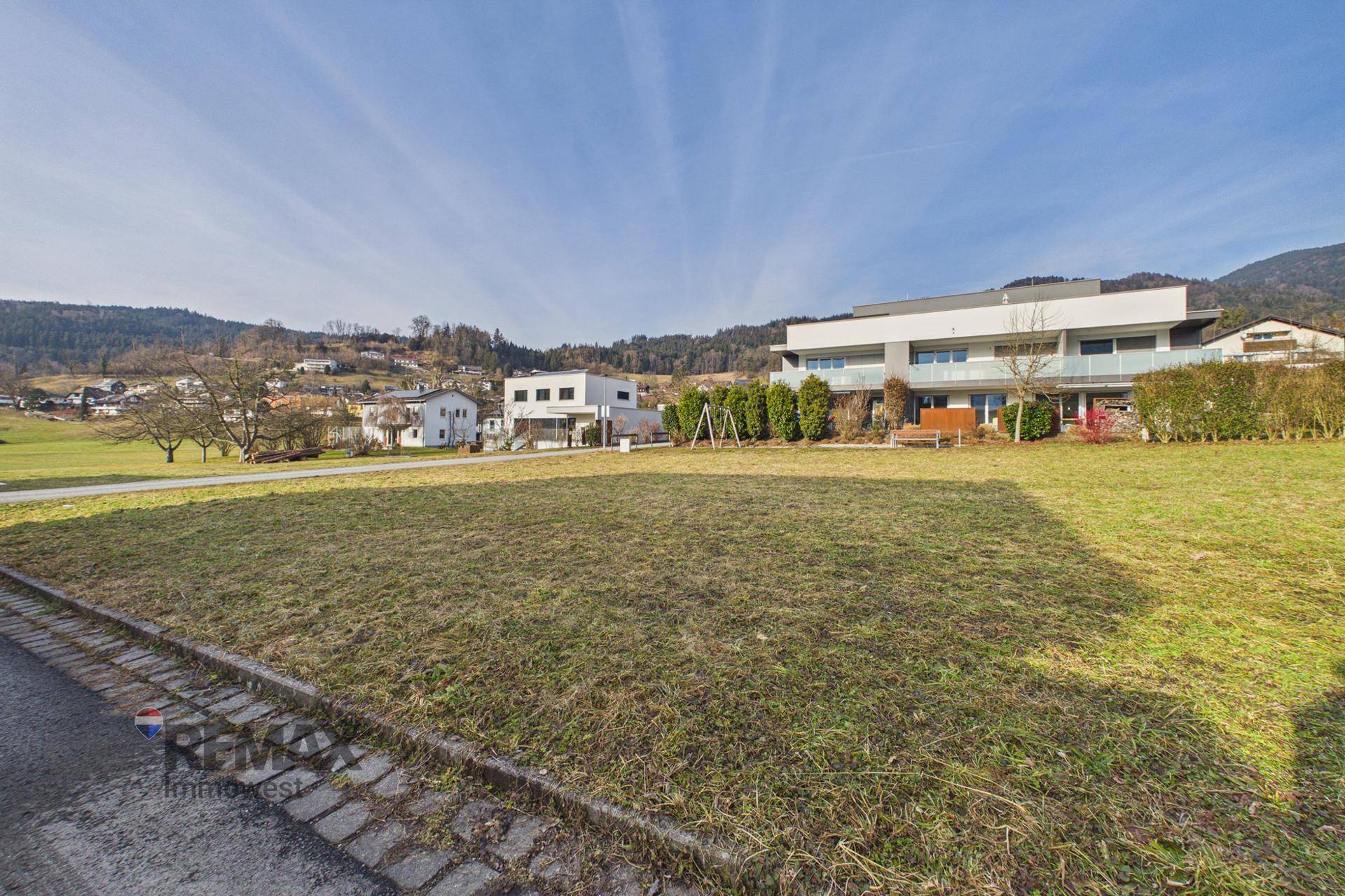 Blick über eine grüne Wiese auf moderne Häuser und bewaldete Hügel im Hintergrund.