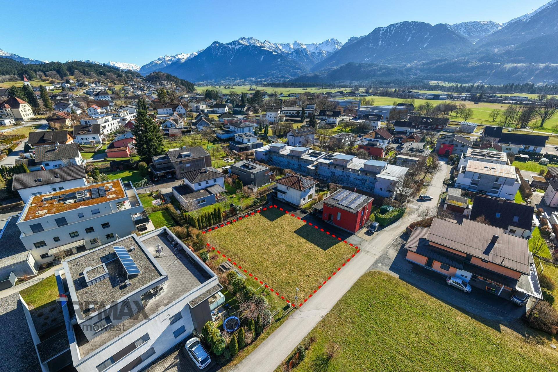 Luftaufnahme eines Wohngebiets mit markiertem Grundstück, umgeben von grünen Feldern und Bergen.