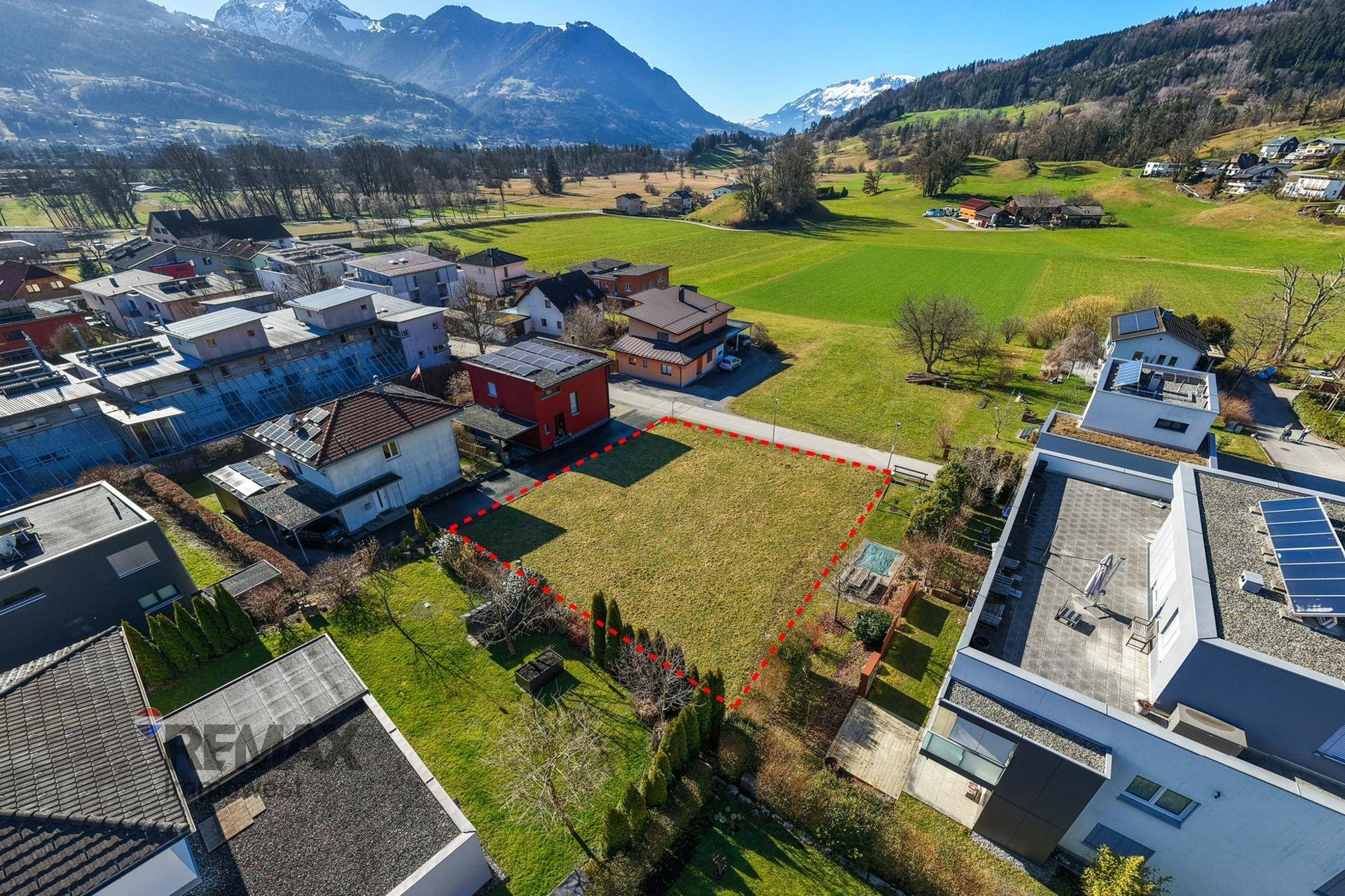 Weitwinkel-Luftaufnahme eines Wohngebiets mit markiertem Grundstück, umgeben von grünen Feldern und Bergen.