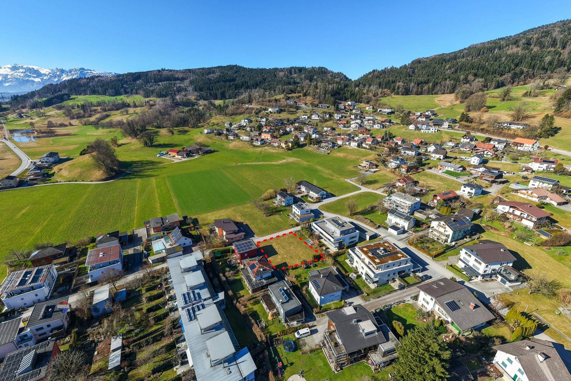 Luftaufnahme eines Wohngebiets mit markiertem Grundstück, umgeben von grünen Feldern, Bergen und einem Teich.