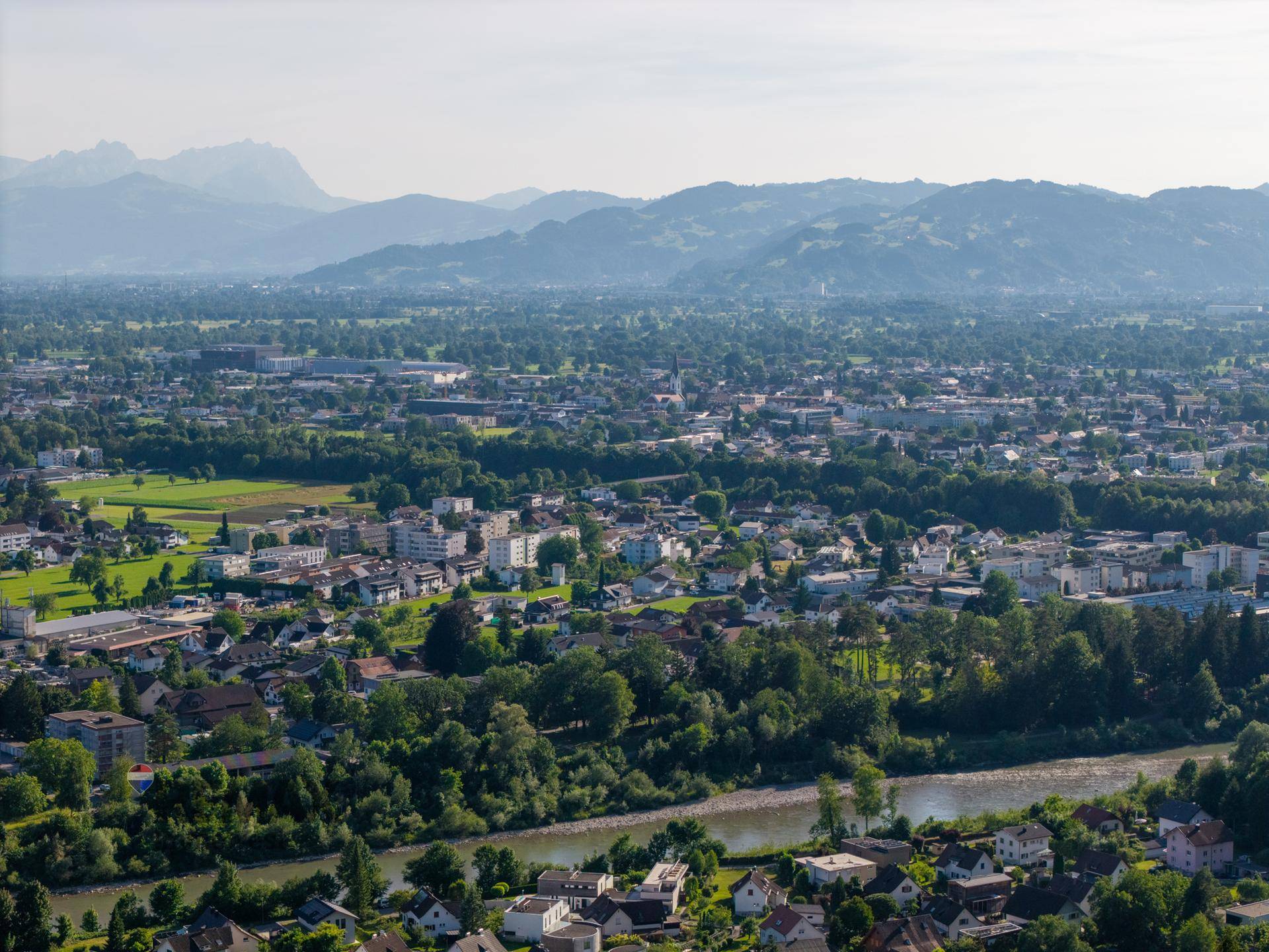 Weitläufiger Blick über die Stadt und die umliegende grüne Landschaft mit Bergen im Hintergrund.
