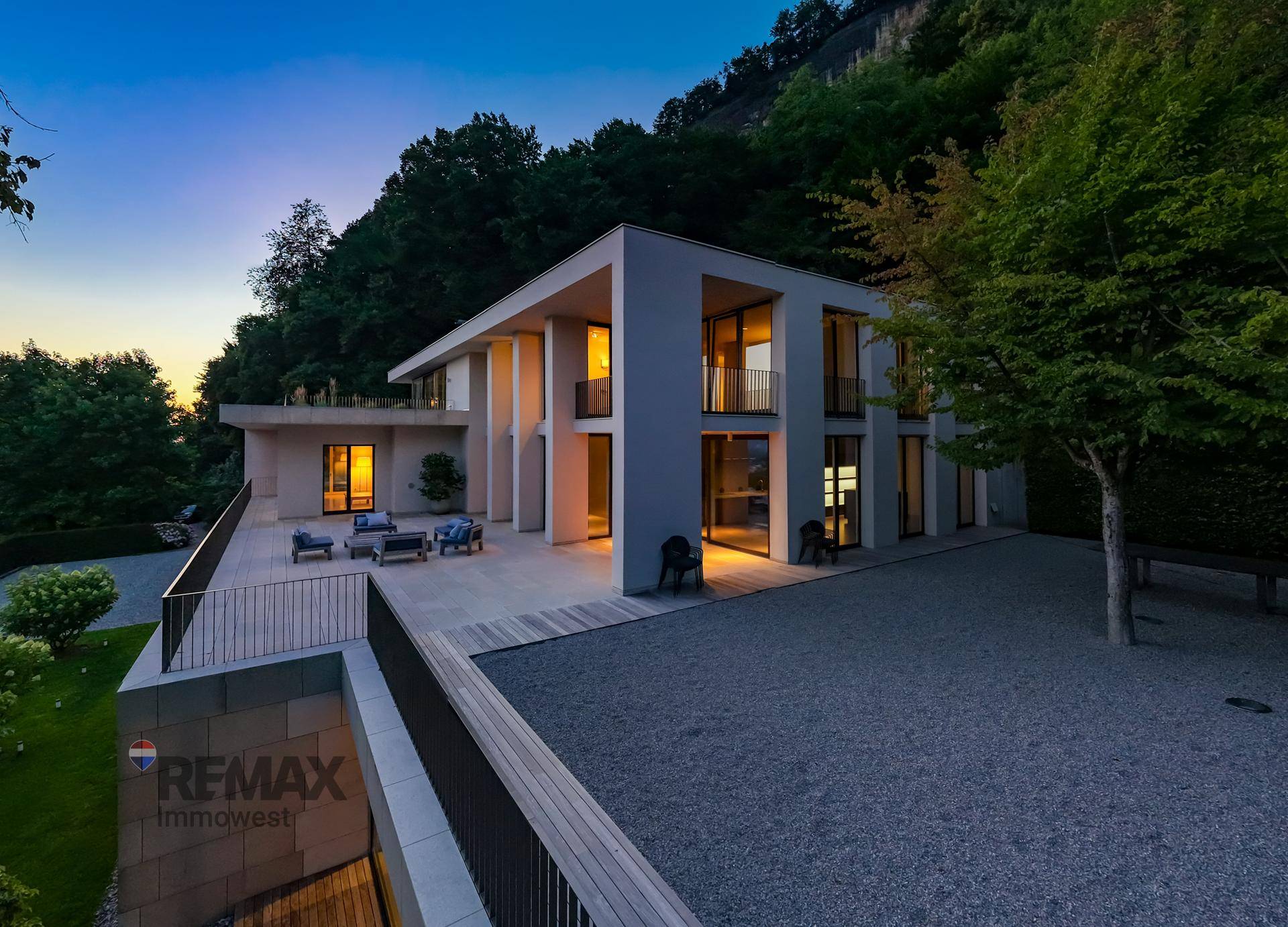 Beleuchtete Terrasse der modernen Villa in der Abenddämmerung, mit Loungemöbeln und Kiesfläche.