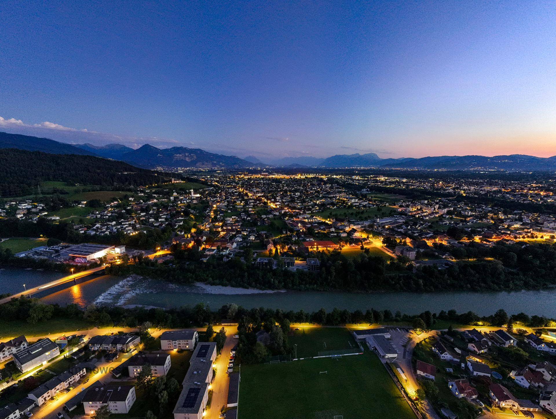 Nächtlicher Panoramablick über die beleuchtete Stadt und die umliegende Landschaft mit Fluss und Bergen.