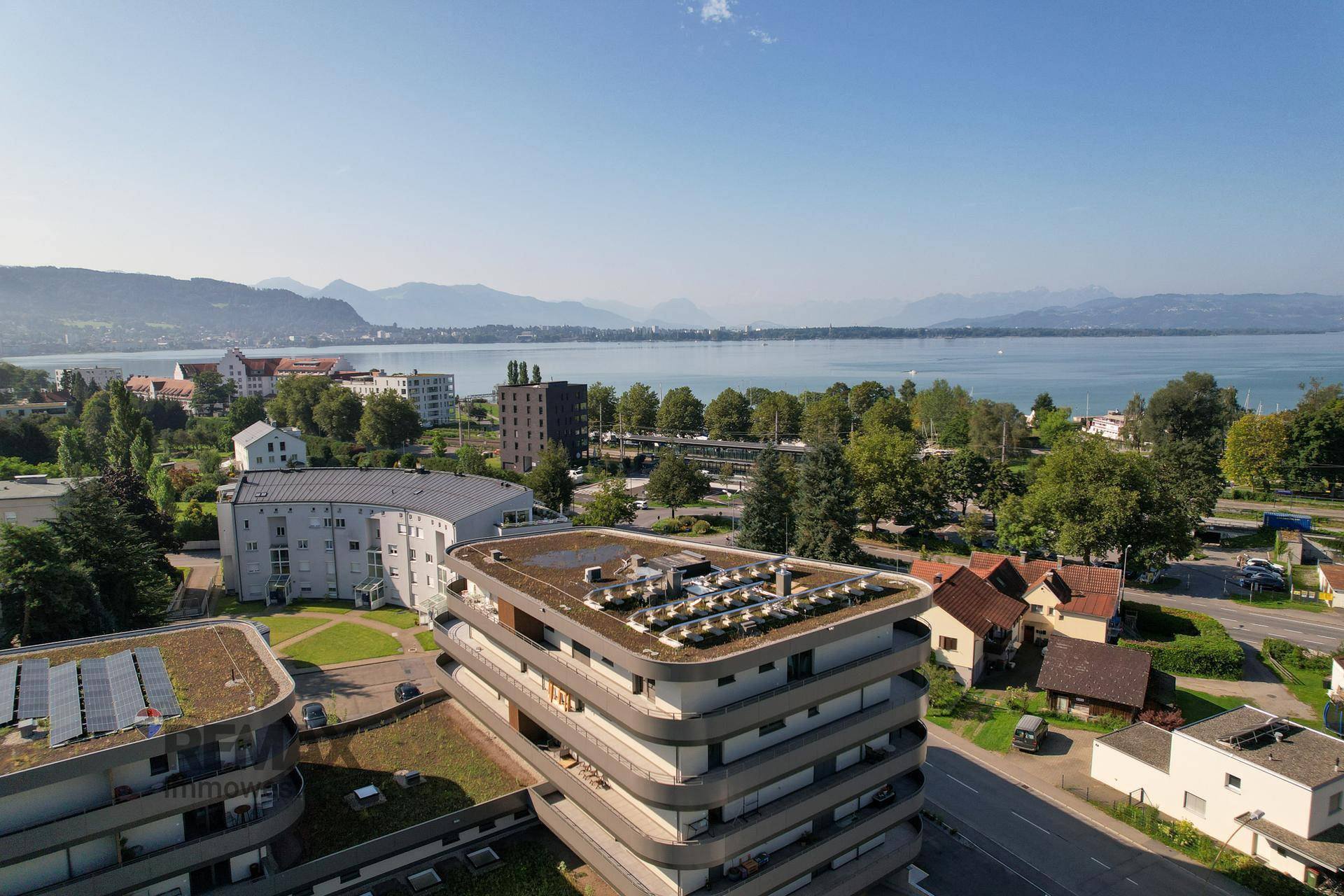Luftaufnahme eines modernen Gebäudes mit Gründach, Solaranlagen und weitem Blick auf den See und die Berge.