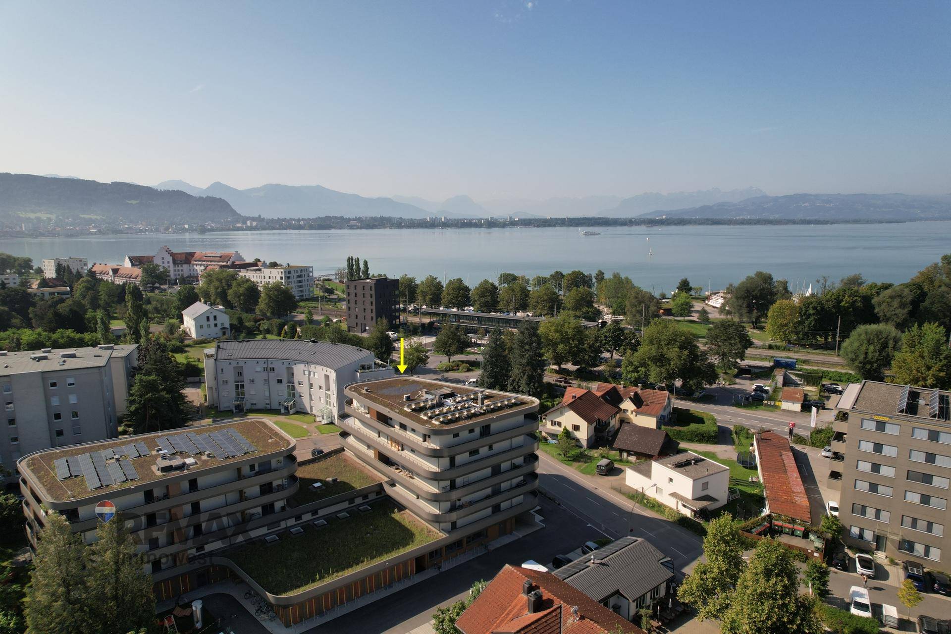 Luftaufnahme eines modernen Gebäudes mit Gründach, Solaranlagen und weitem Blick auf den See und die Berge.