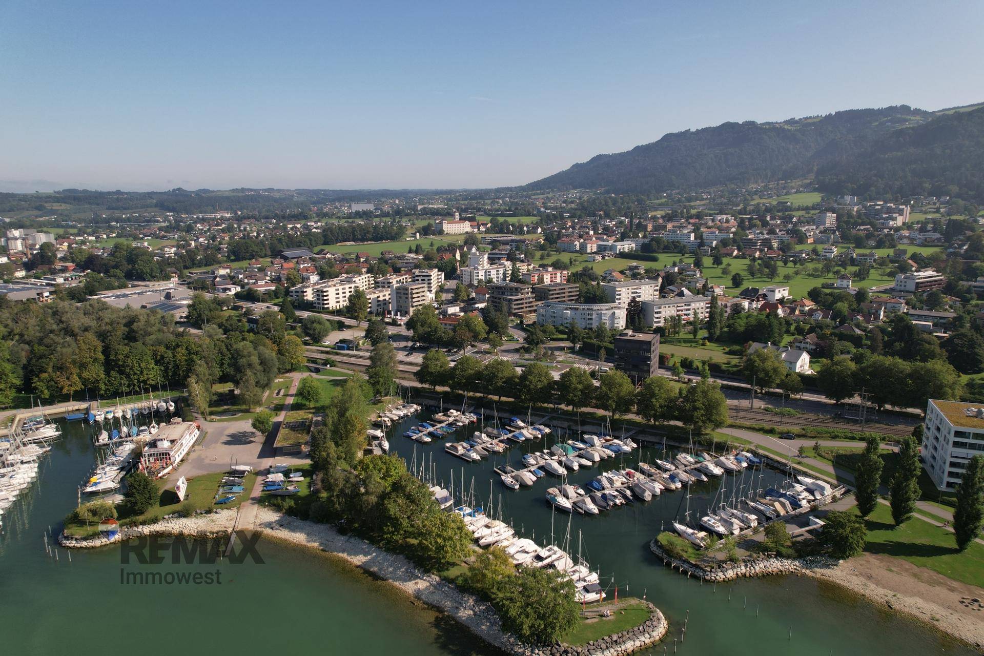 Luftaufnahme einer Stadt mit Yachthafen, See und Bergen im Hintergrund unter blauem Himmel.