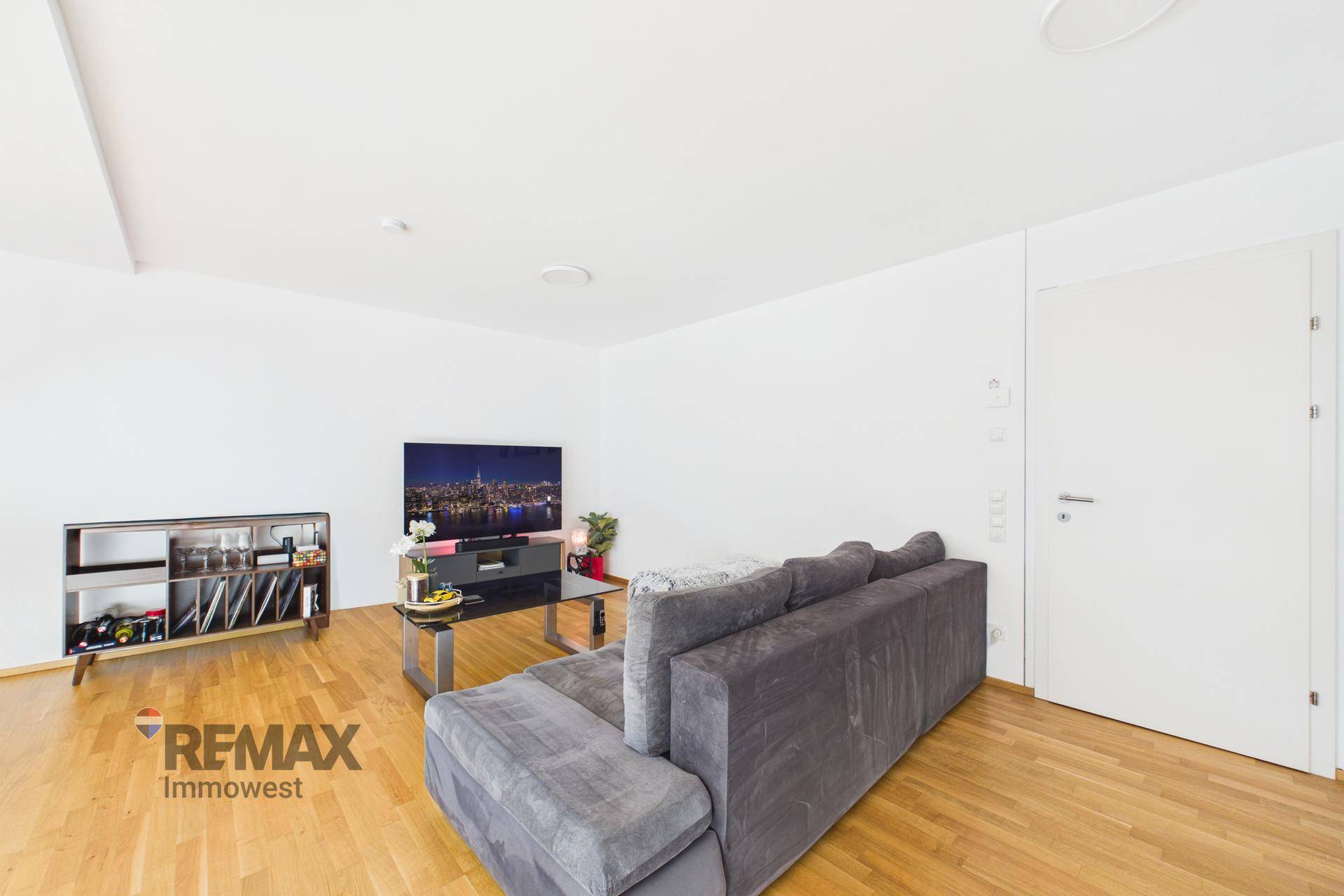 Gemütliches Wohnzimmer mit Sofa, Fernseher und Holzfußboden in heller Atmosphäre.