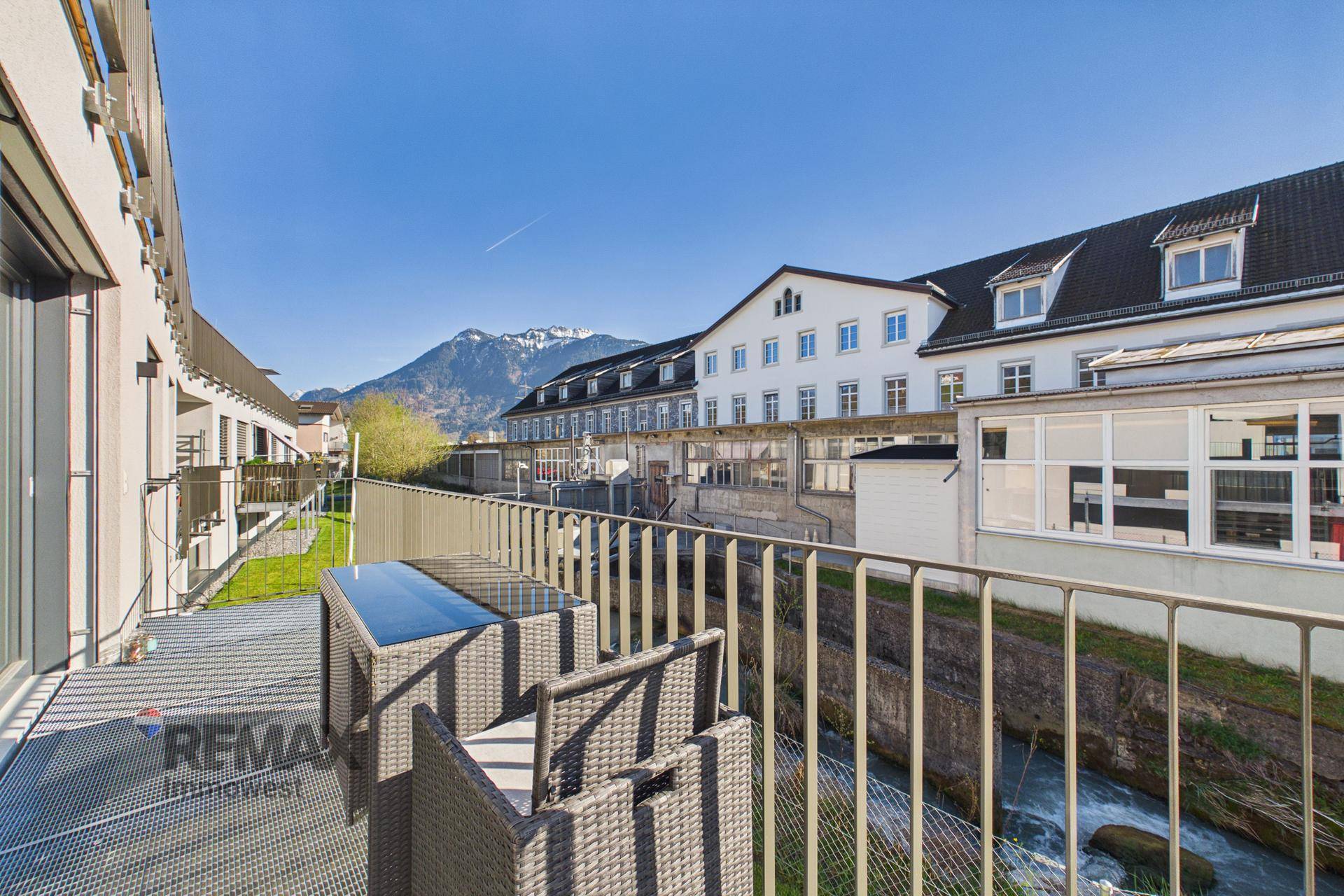 Sonniger Balkon mit Sitzgelegenheiten und Blick auf die Berge und einen Bach.