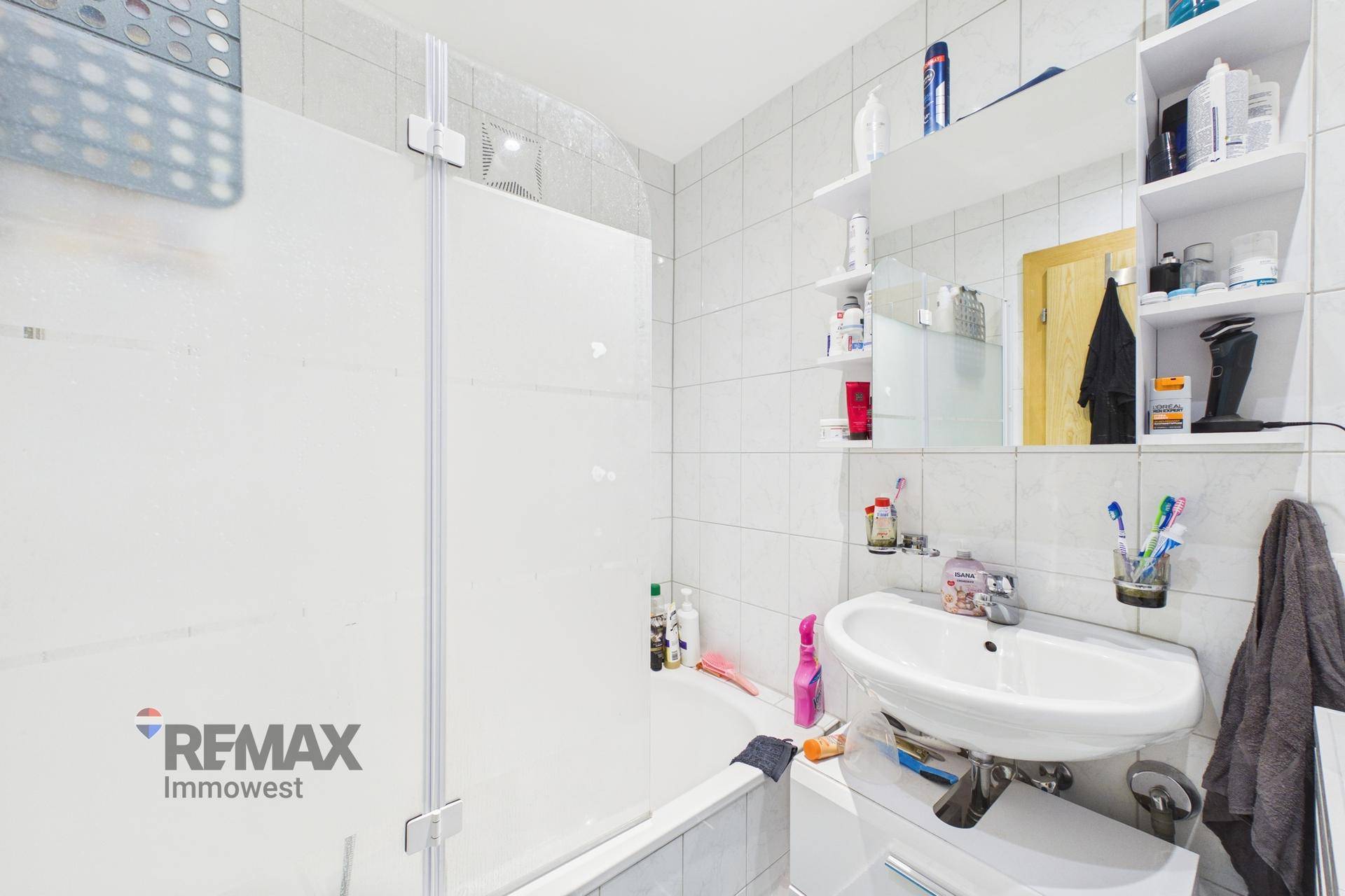 Badezimmer mit weißen Fliesen, Badewanne mit Duschwand, Waschbecken und Spiegelschrank.