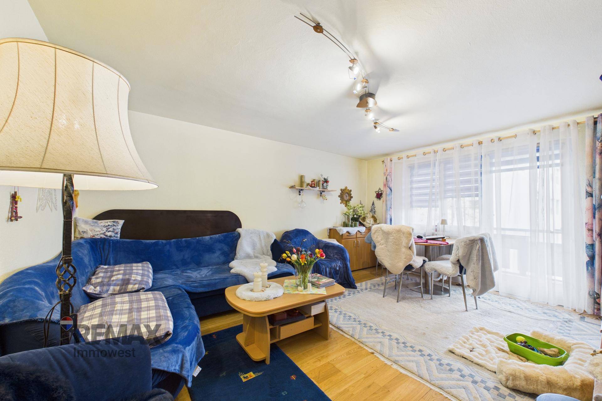 Gemütliches Wohnzimmer mit blauer Couch, Holzboden und einem Esstischbereich am Fenster.