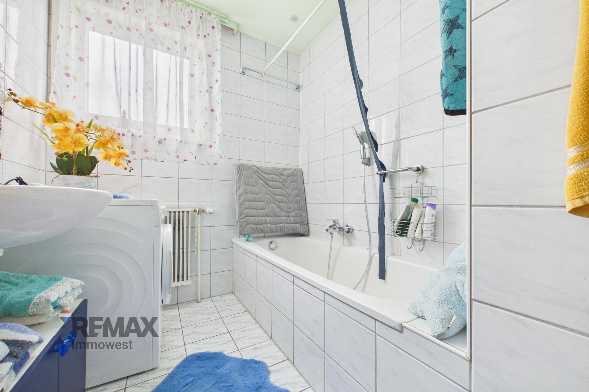 Badezimmer mit weißen Fliesen, Badewanne mit Duschfunktion und einem Fenster.