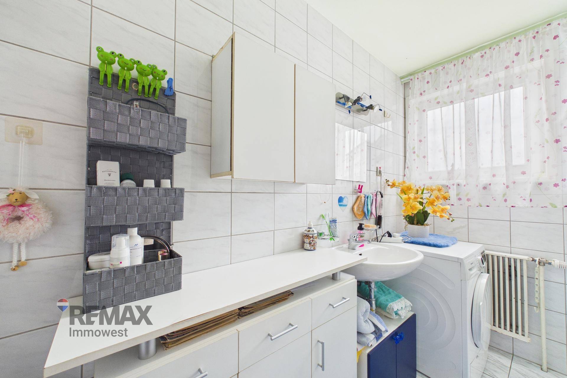 Badezimmer mit weißen Fliesen, Waschbecken, Waschmaschine und einem Fenster.