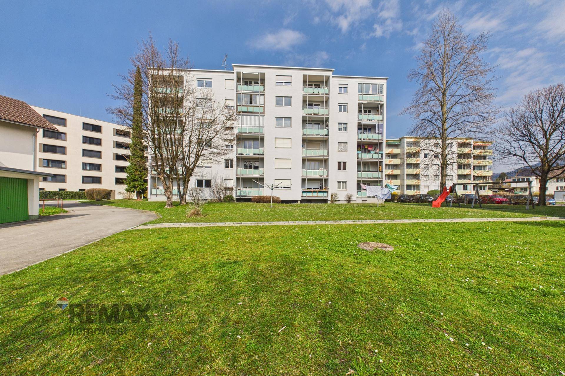 Wohngebäude mit grüner Rasenfläche und Spielplatz im Vordergrund unter blauem Himmel.