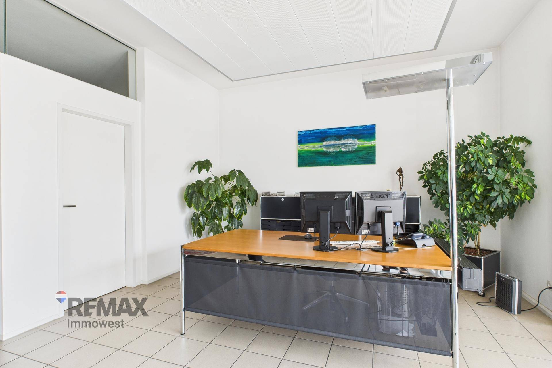 Modernes Büro mit großem Schreibtisch, mehreren Monitoren, Pflanzen und hellem Fliesenboden.