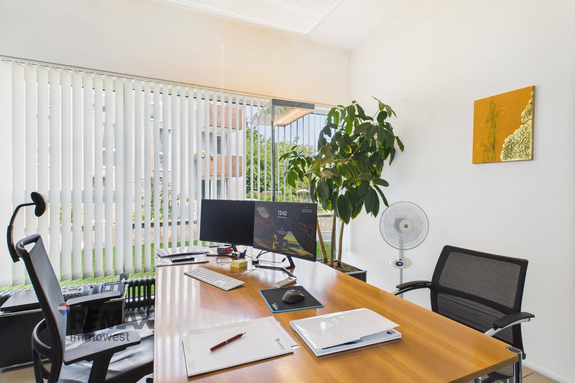 Privates Büro mit Schreibtisch, Computermonitoren und vertikalen Jalousien an den Fenstern.