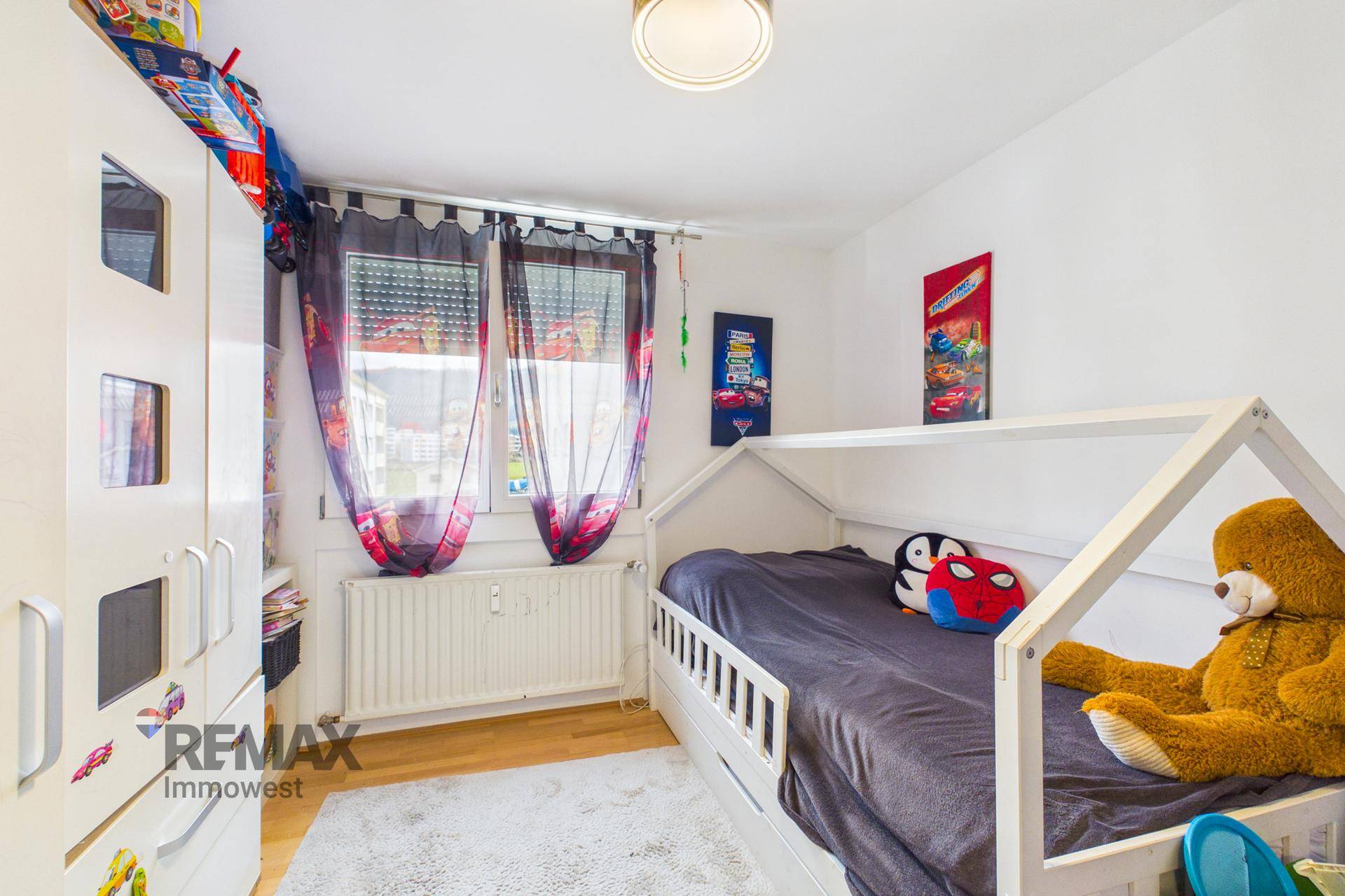 Kinderzimmer mit hellem Holzfußboden, Fenster und einem gemütlichen Hausbett.