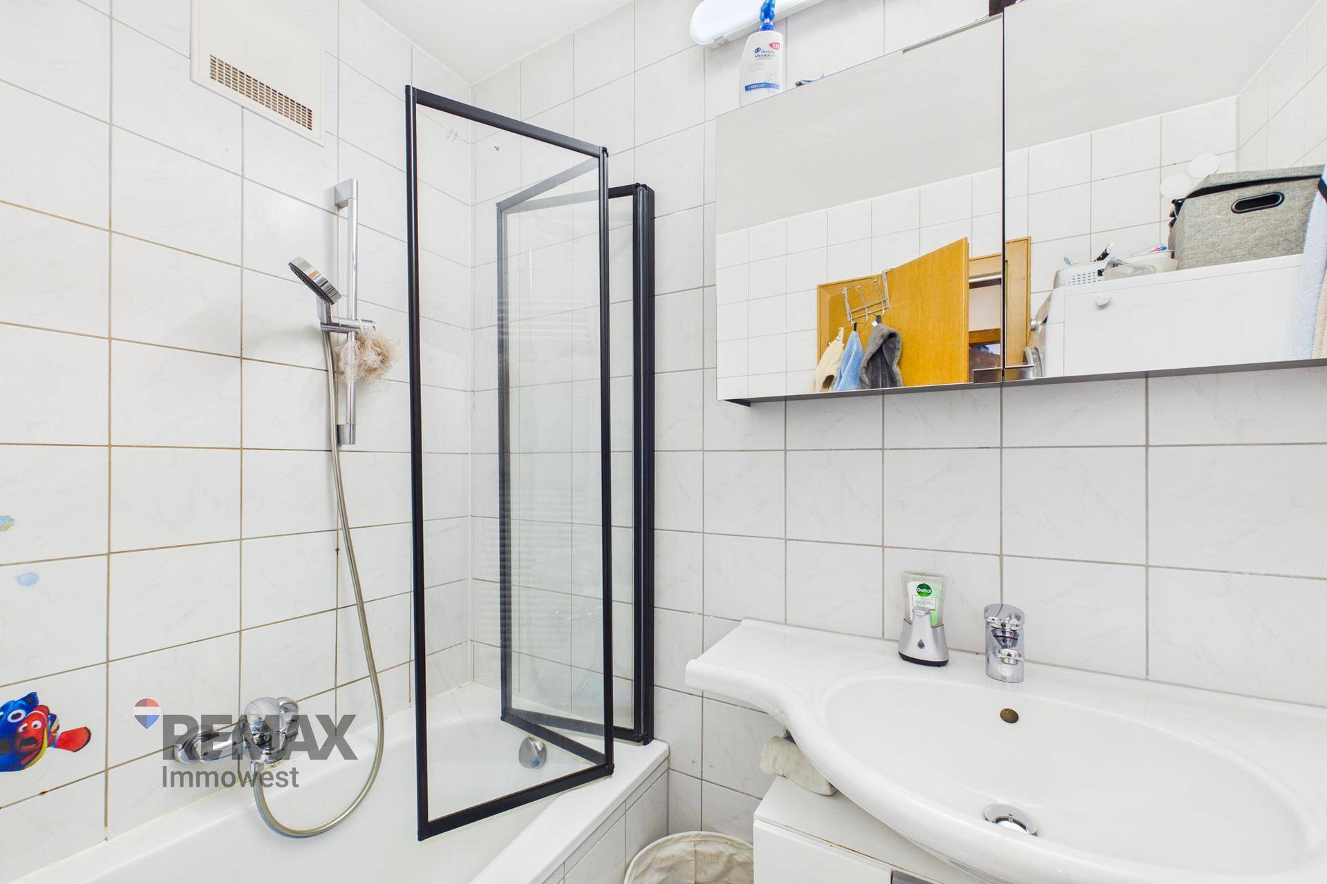 Helles Badezimmer mit weißen Fliesen, Waschbecken und Badewanne mit Duschabtrennung.