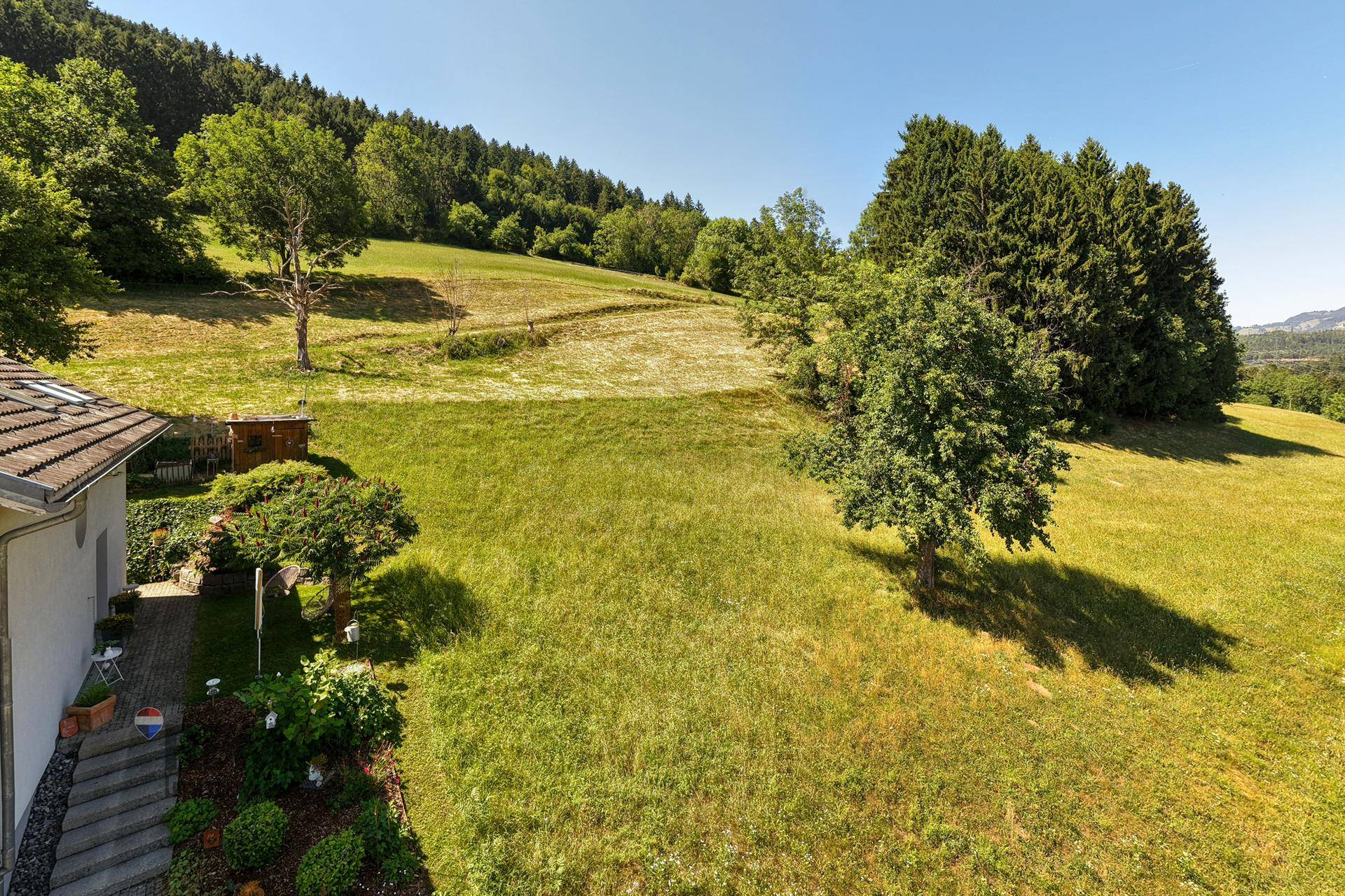 Seitlicher Blick auf ein Haus mit gepflegtem Garten und angrenzender grüner Wiese am Hang.