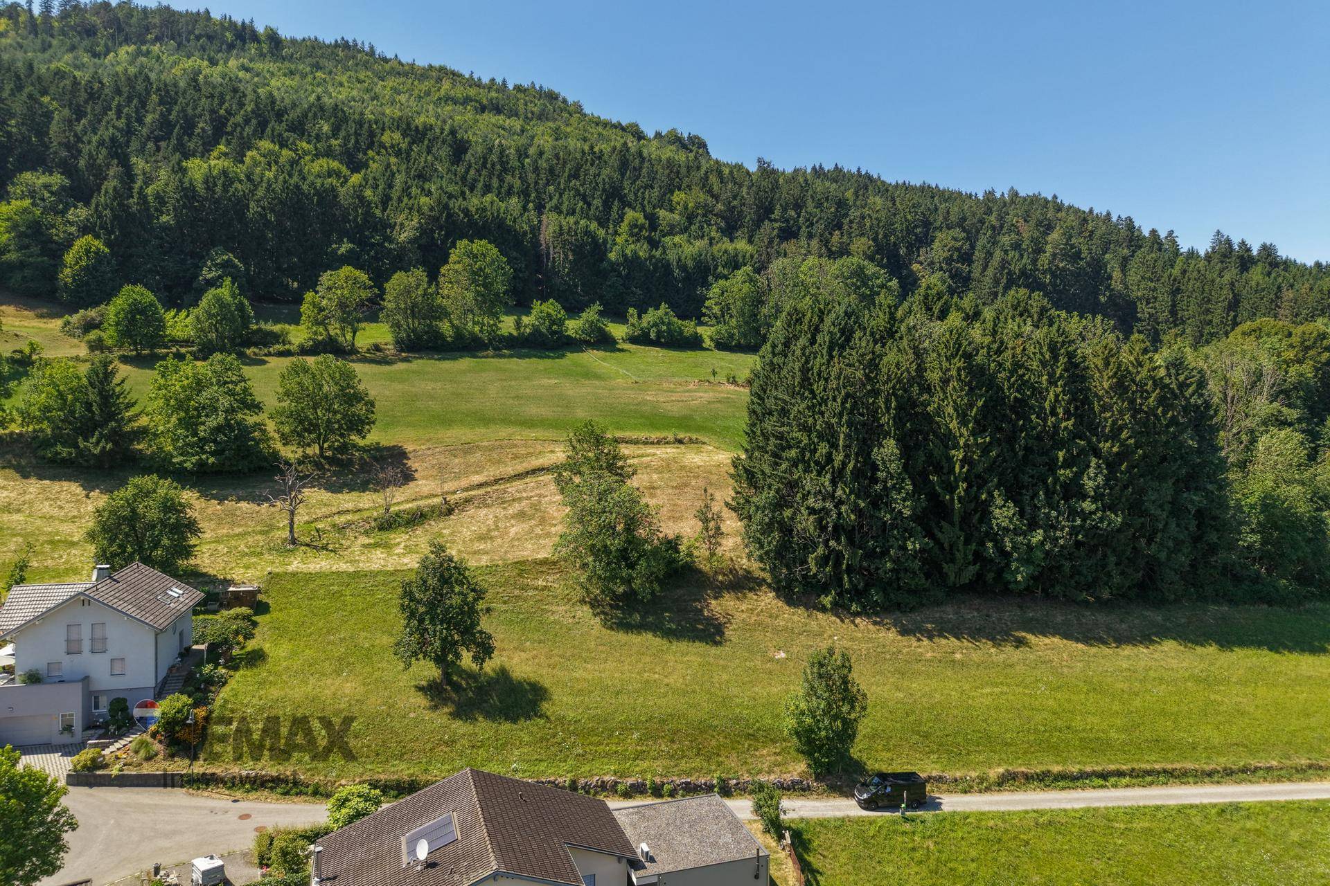 Luftaufnahme von Häusern am Waldrand, umgeben von grünen Wiesen und einem bewaldeten Hügel.