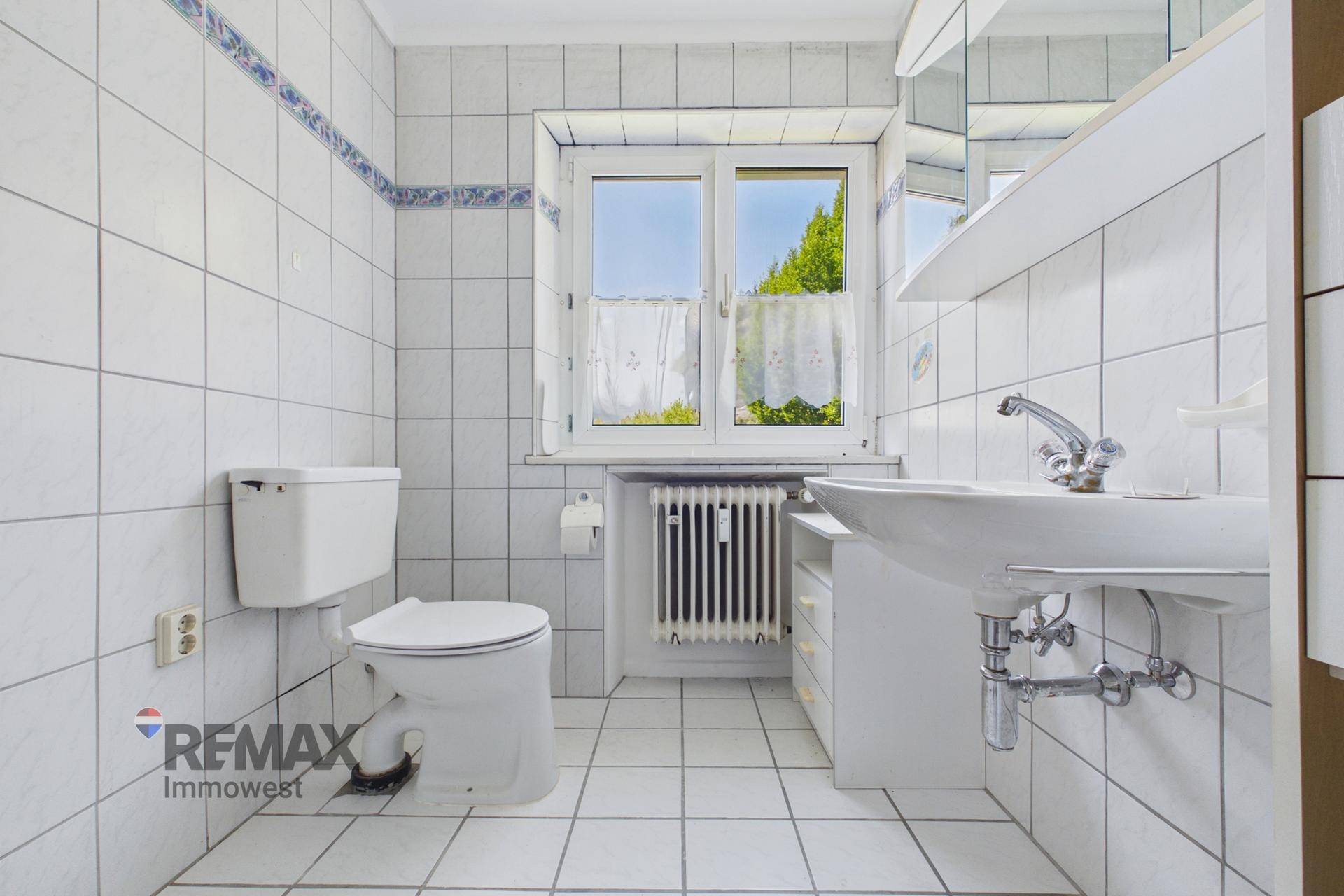 Helles Badezimmer mit weißen Fliesen, WC, Waschbecken und einem Fenster für Tageslicht.