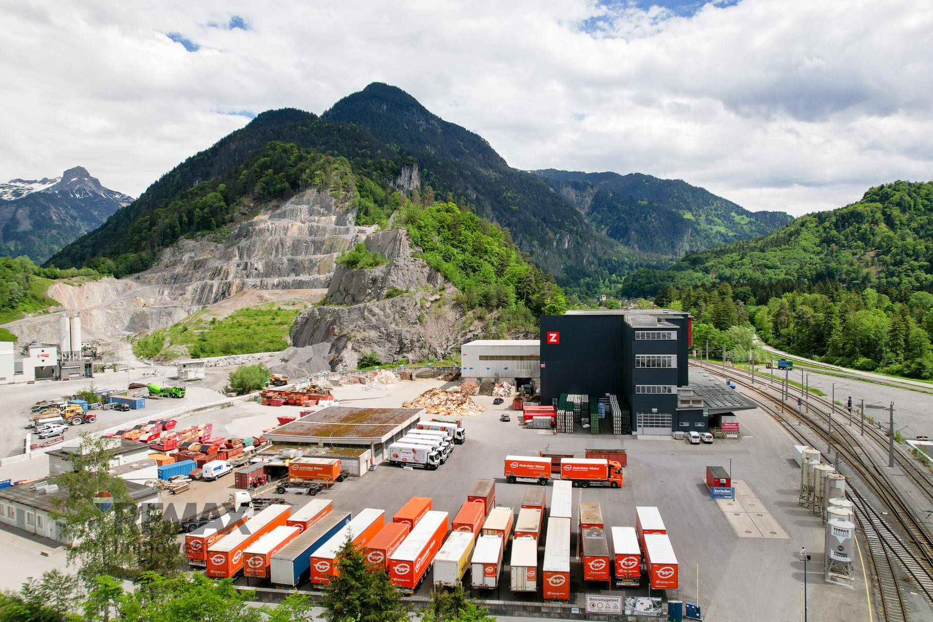 Weitläufiges Industriegebiet mit Lagerflächen und Transportfahrzeugen, umgeben von grünen Hügeln und einem Steinbruch.