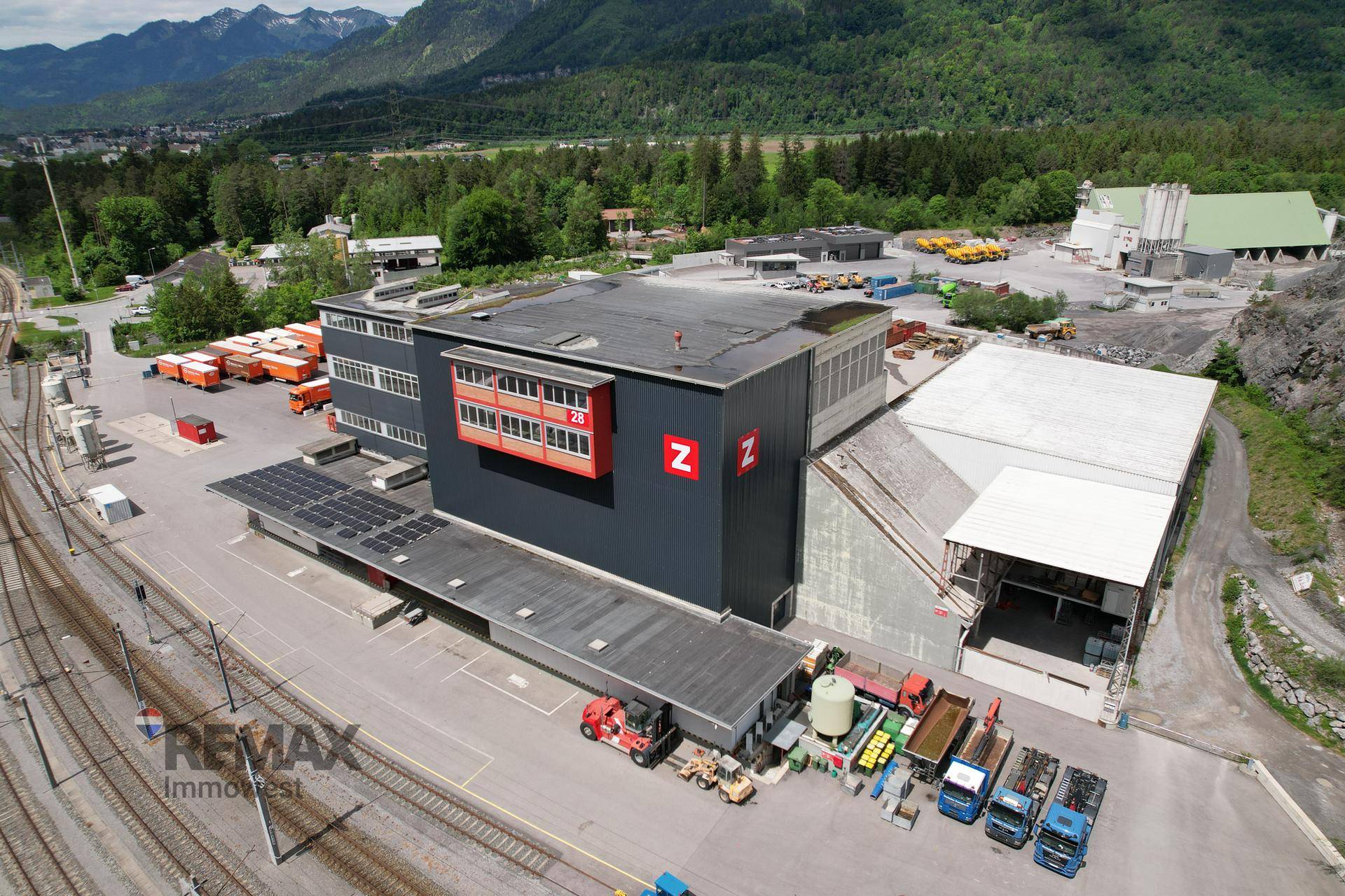 Übersicht über ein großes Industriegelände mit mehreren Gebäuden, Ladezonen und Schieneninfrastruktur vor alpiner Kulisse.