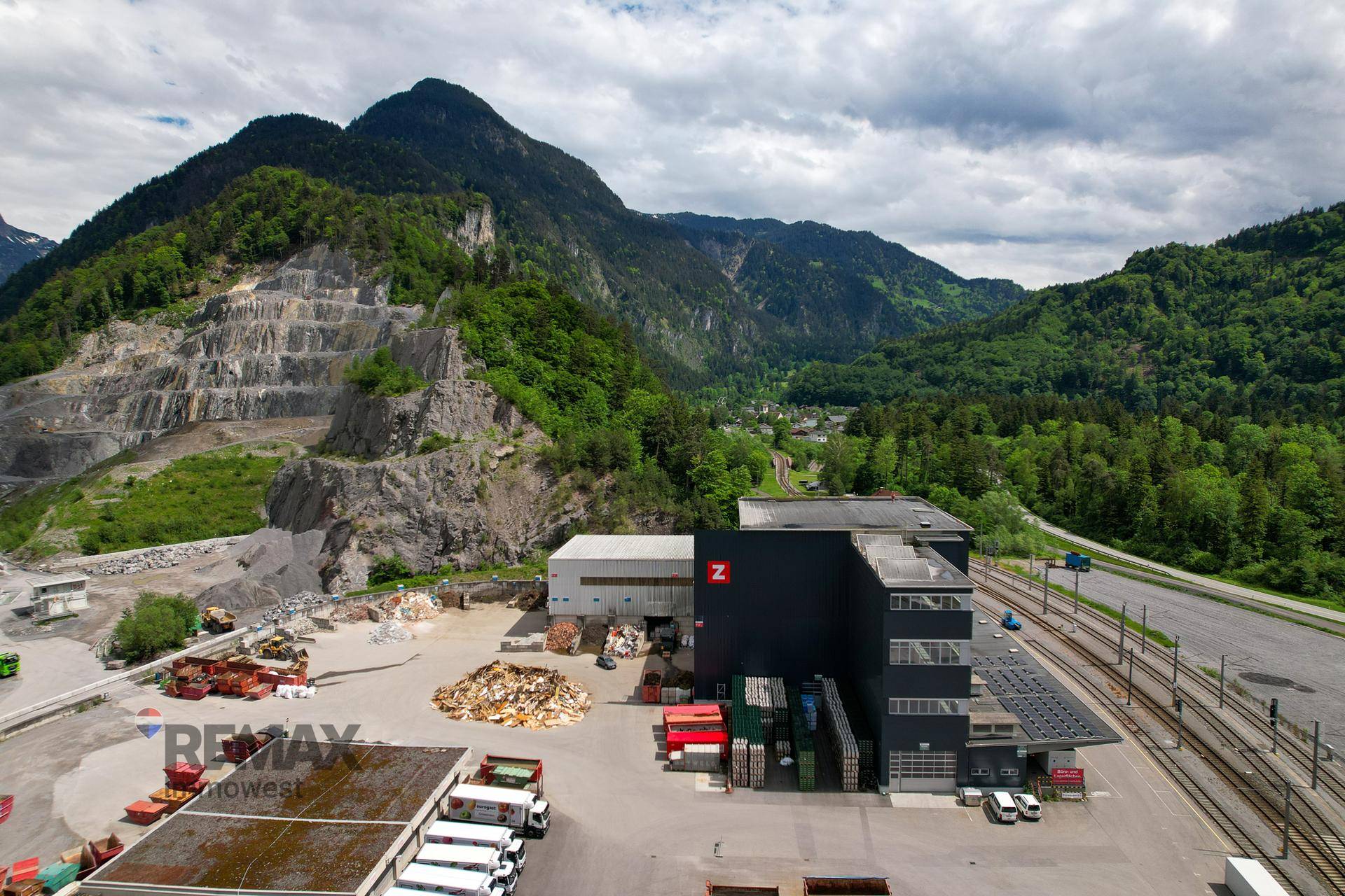 Industriekomplex mit Lagerflächen und Schienenanbindung, eingebettet in eine grüne Berglandschaft mit Steinbruch.