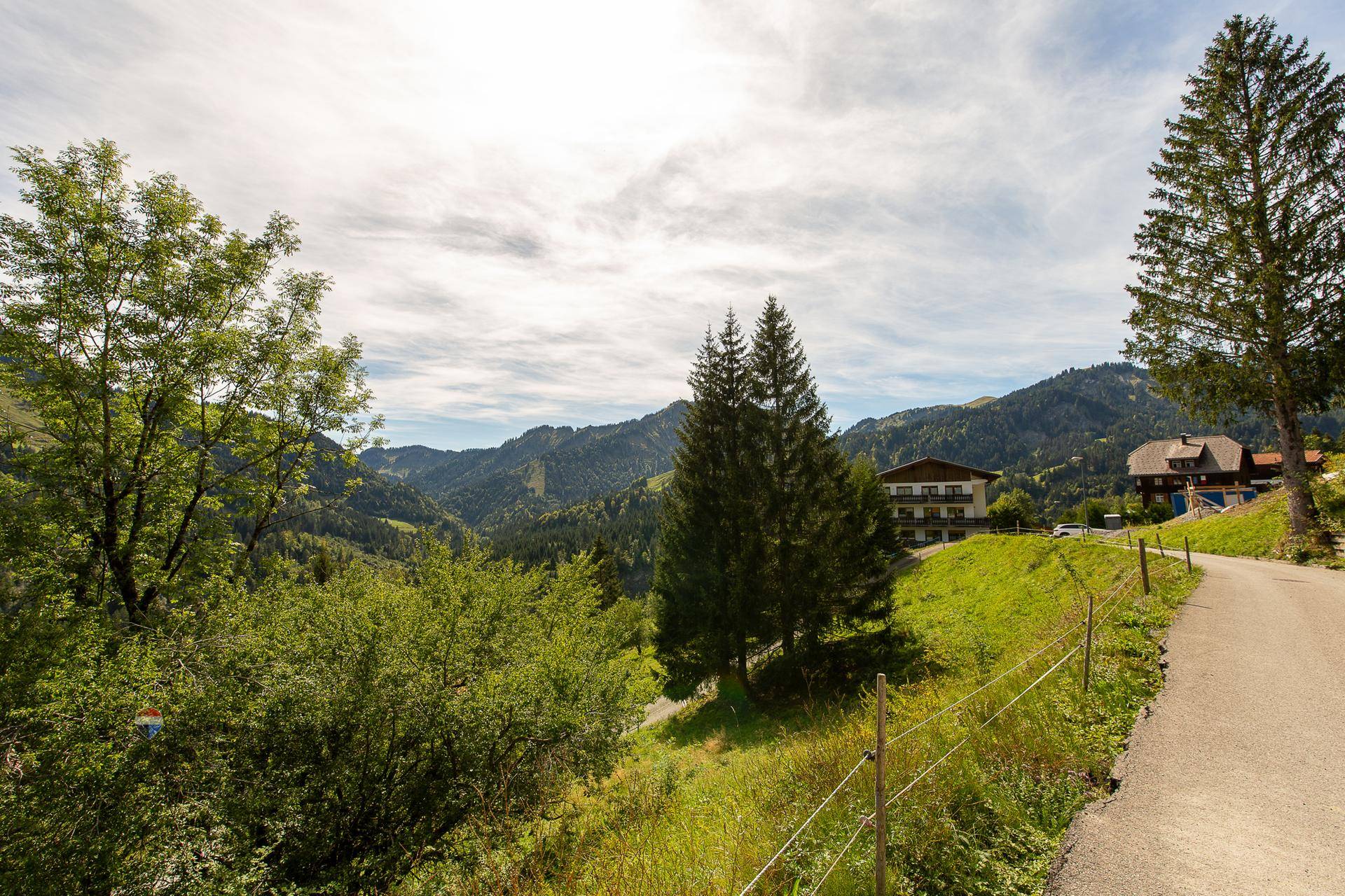 Panoramablick auf eine grüne Berglandschaft mit Häusern und einem Weg unter bewölktem Himmel.