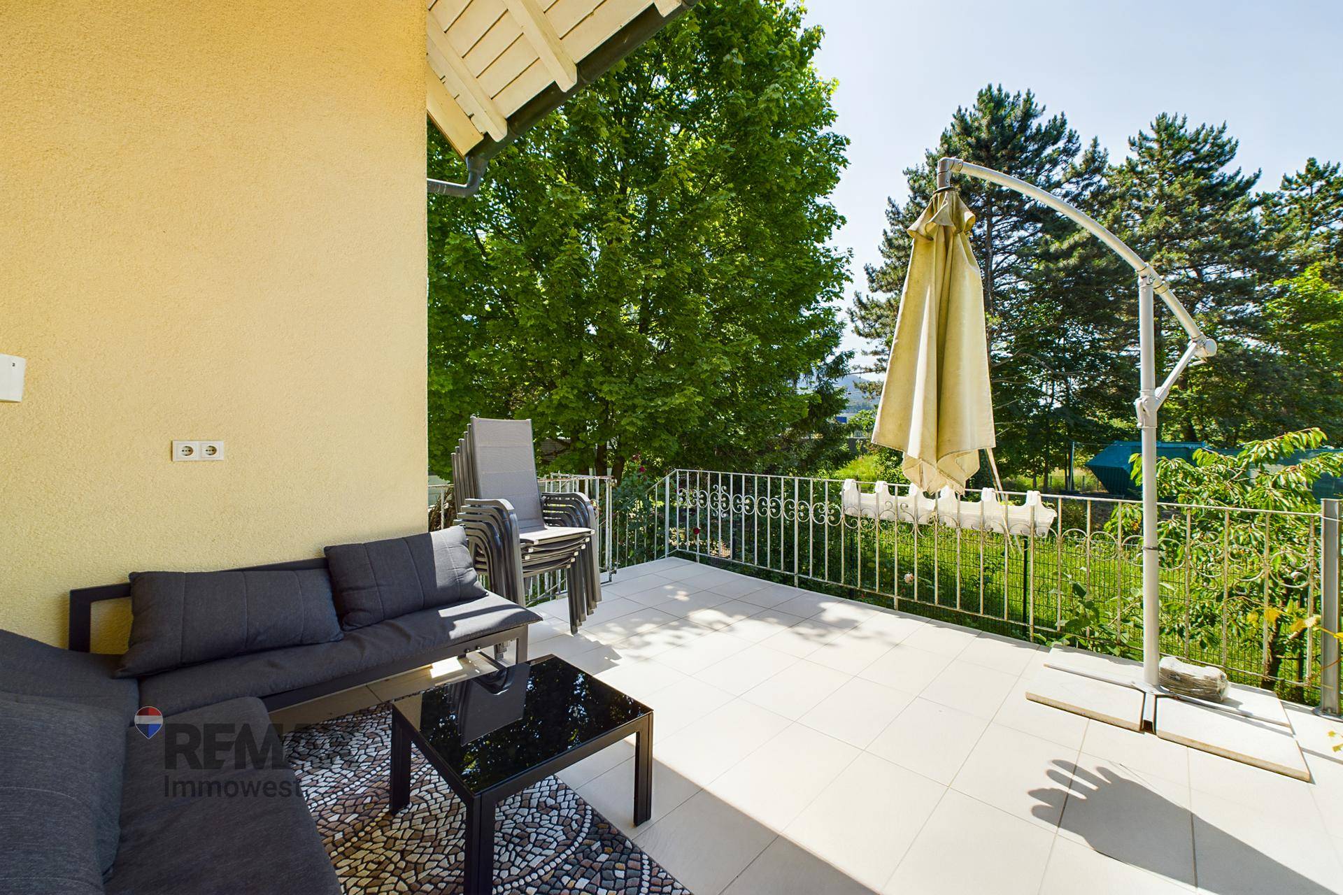 Möblierte Terrasse mit Lounge-Sofa, Sonnenschirm und Blick ins Grüne an einem sonnigen Tag.