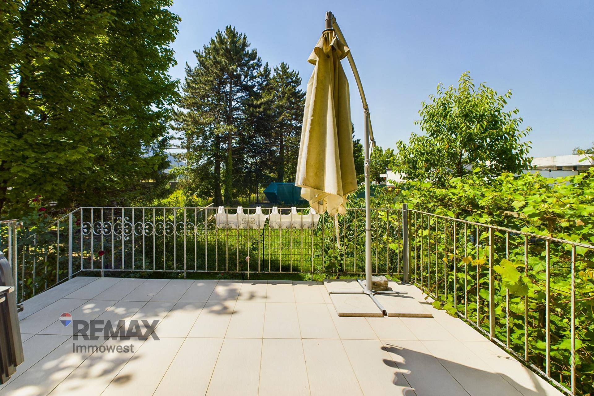 Helle Terrasse mit Fliesenboden, großem Sonnenschirm und schmiedeeisernem Geländer mit Blick ins Grüne.