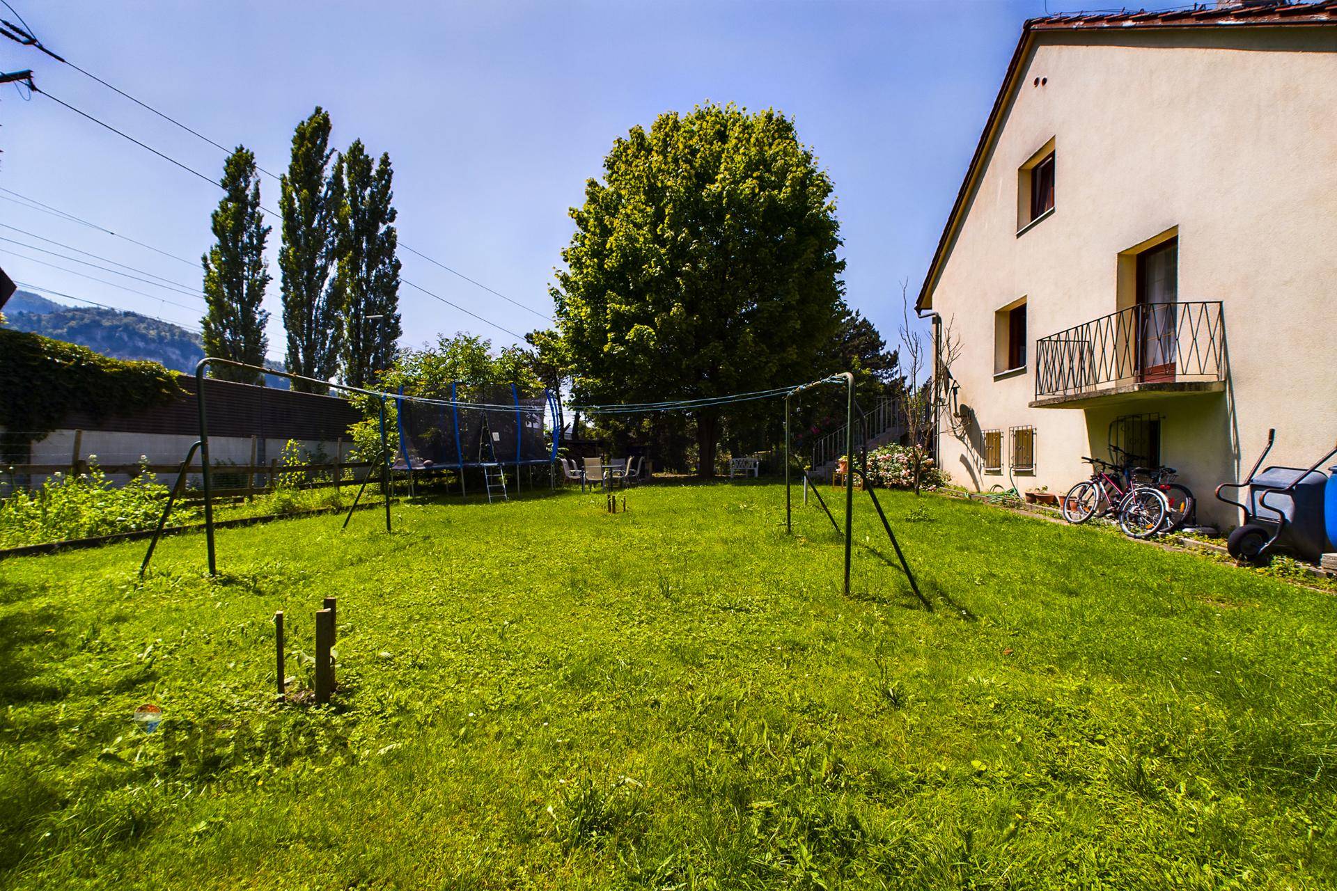 Weitläufiger Garten mit Rasenfläche, Wäscheleine und Trampolin, umgeben von Bäumen und Bergen.