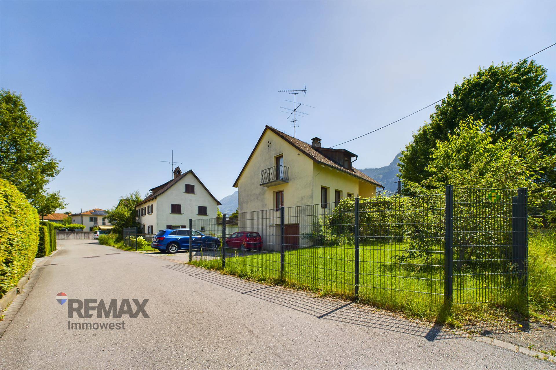 Hausfassade mit Vorgarten, Straße und umliegender Wohnbebauung unter blauem Himmel.