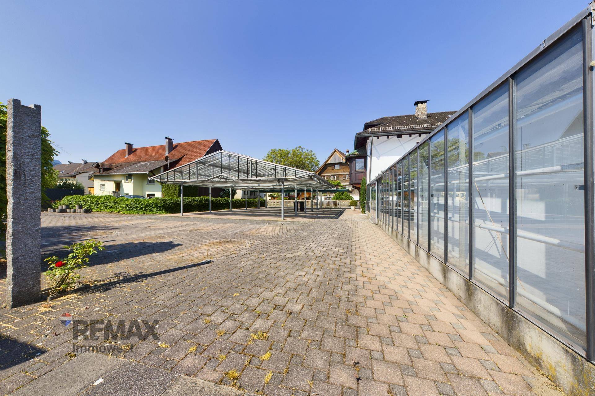 Großer gepflasterter Außenbereich mit Parkplätzen und Blick auf das Gewächshaus und umliegende Häuser.
