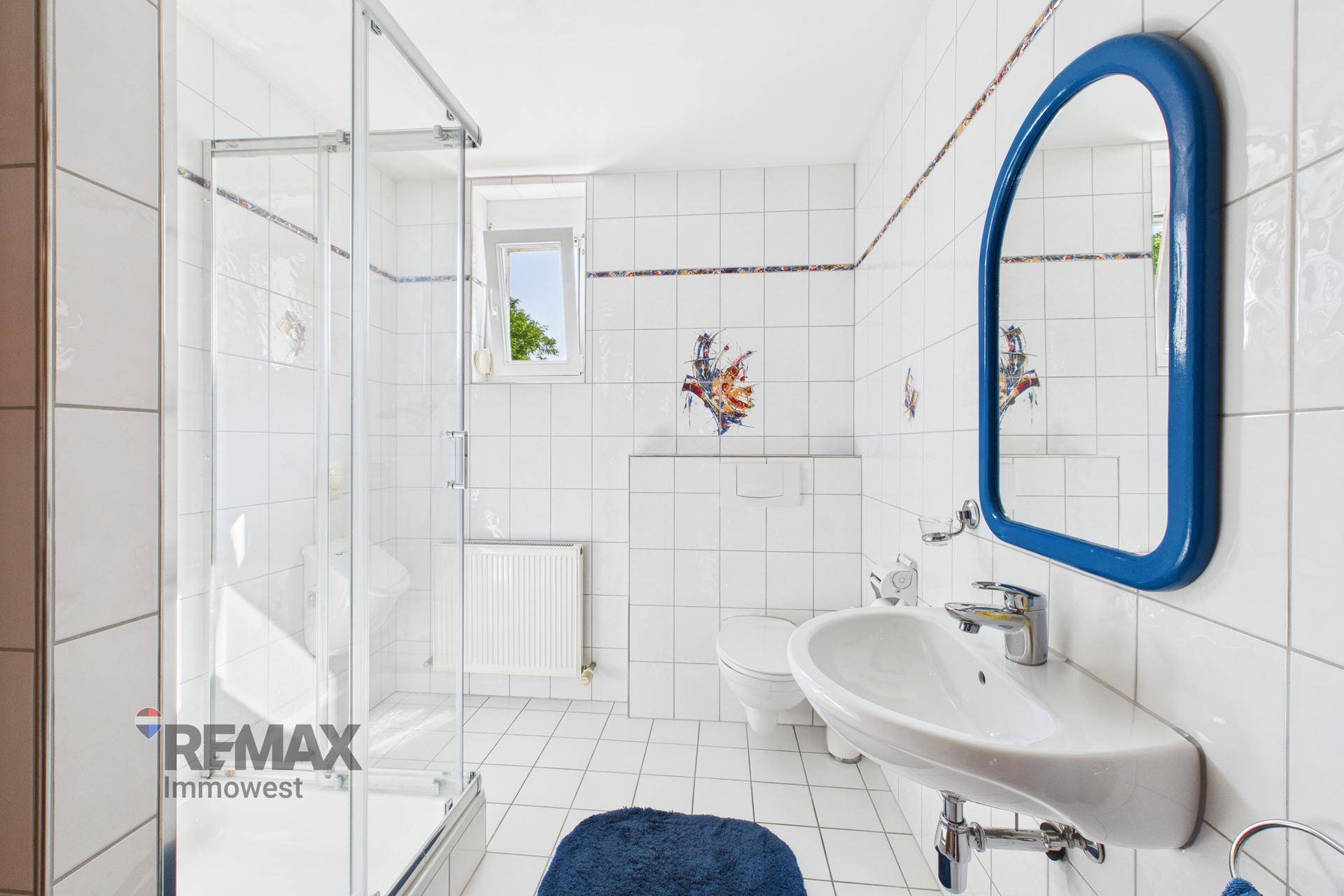 Helles Badezimmer mit weißen Fliesen, Duschkabine, Waschbecken und WC.