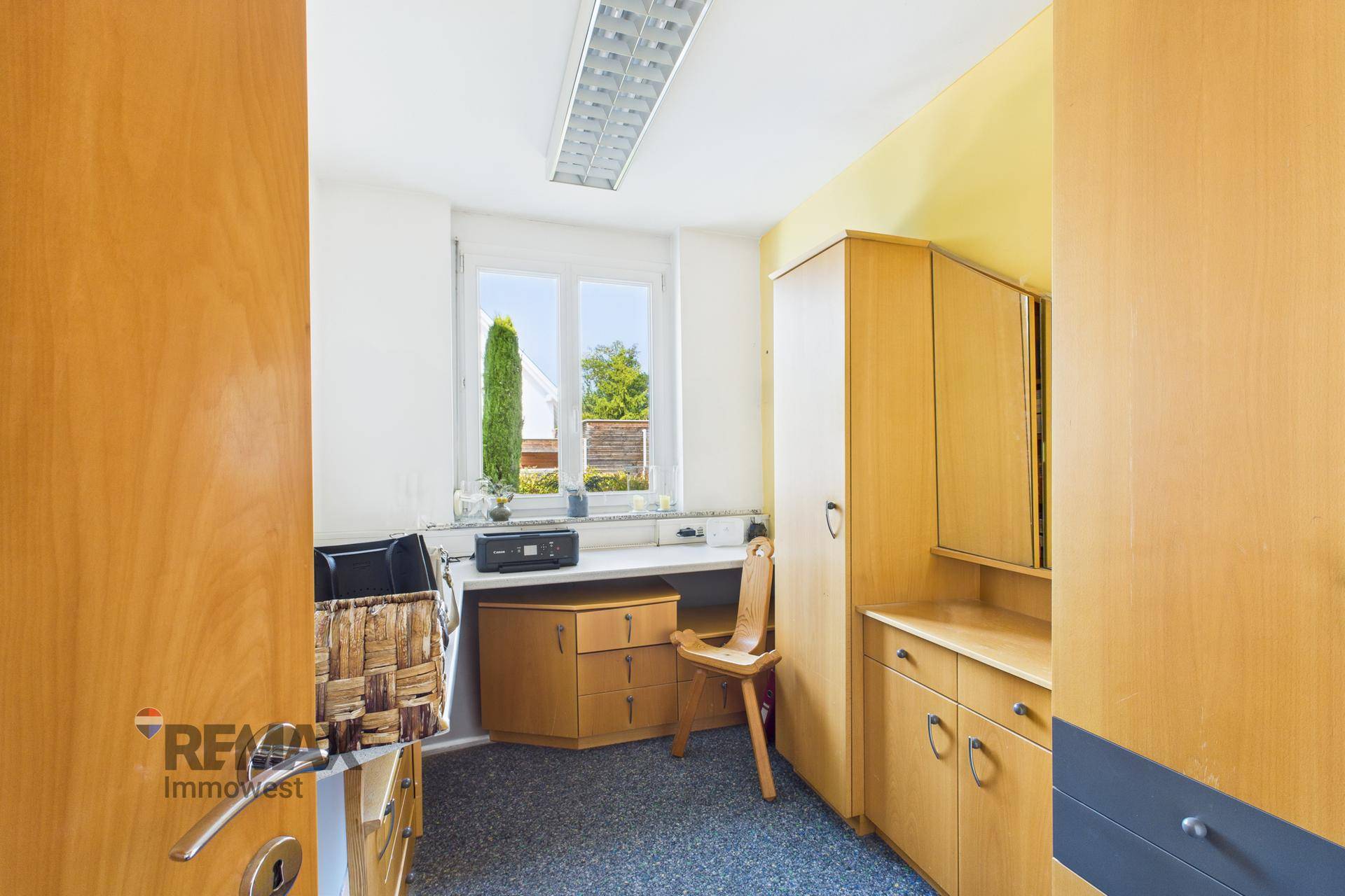 Funktionales Büro mit Holzmöbeln, Teppichboden und Fenster mit Blick ins Grüne.