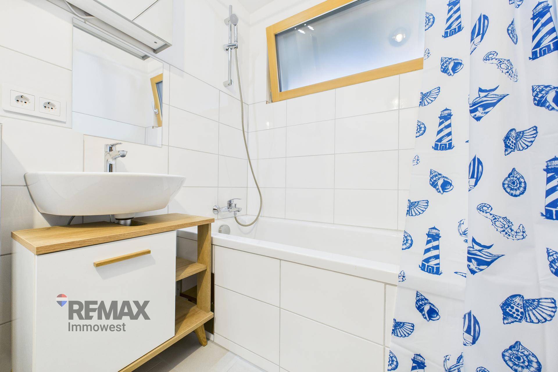 Helles Badezimmer mit Badewanne, Waschbecken, Fenster und maritimem Duschvorhang.