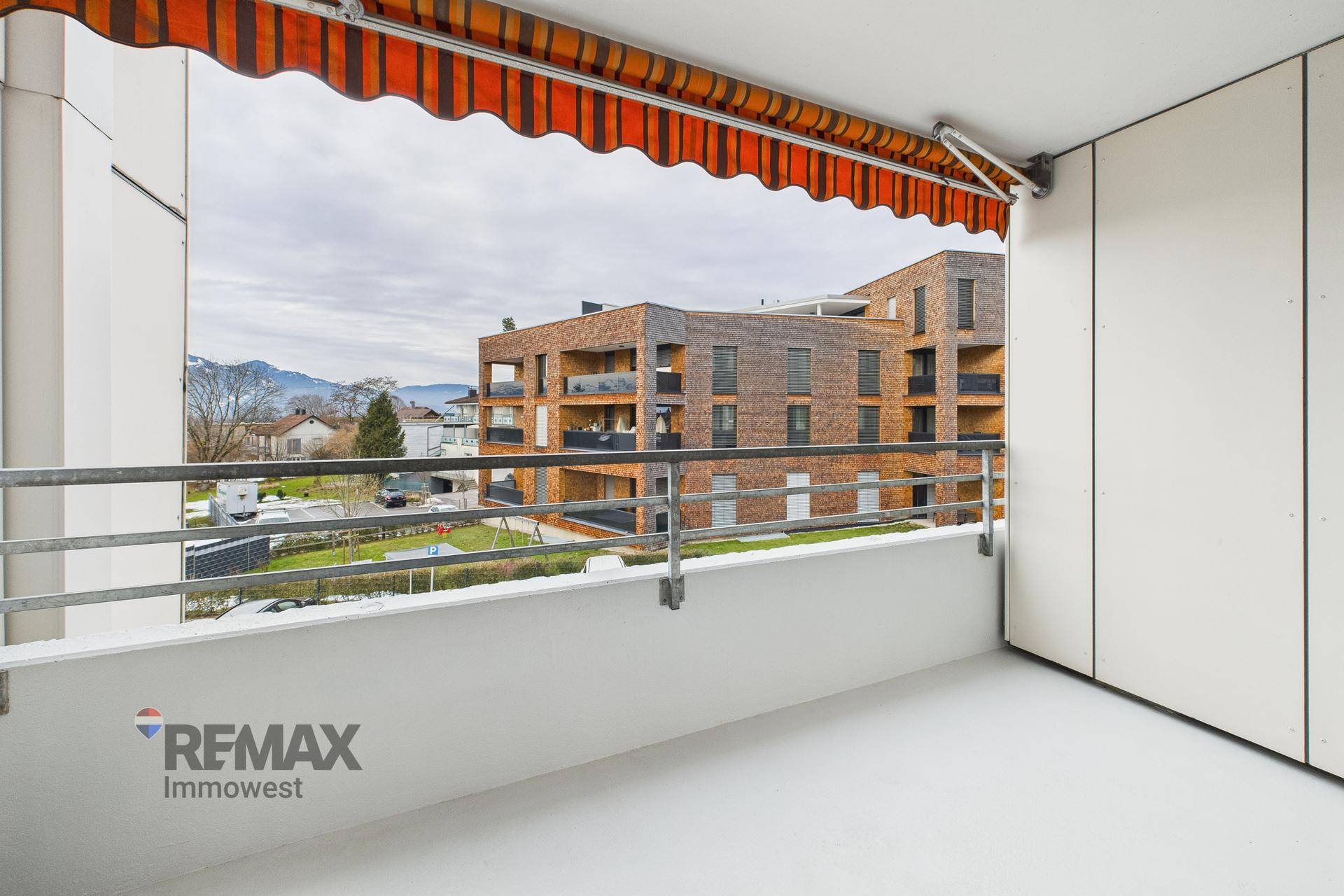 Balkon mit Markise und Blick auf moderne Nachbargebäude und Berge im Hintergrund.