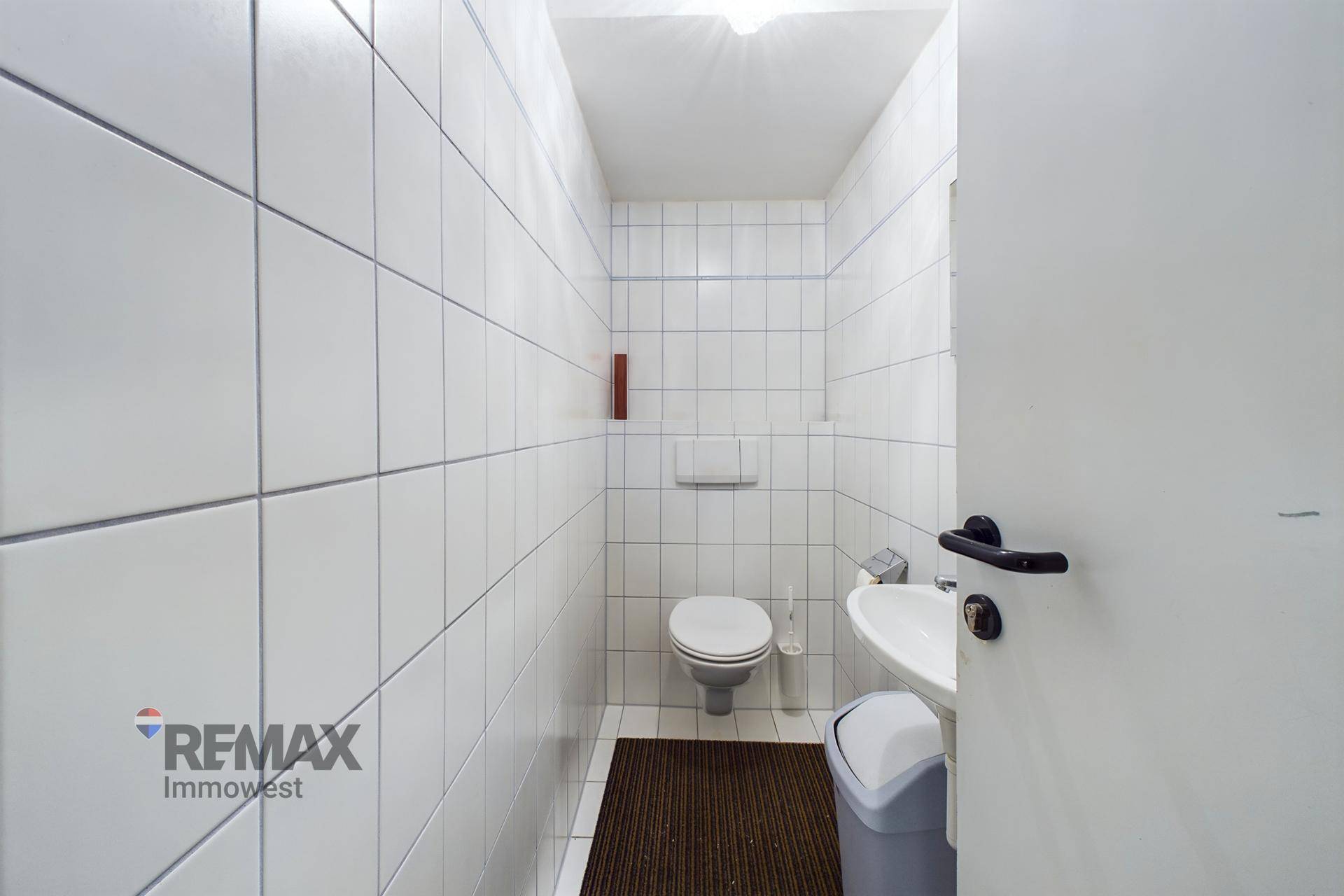 Kleiner, sauberer Toilettenraum mit weißen Fliesen, einer Toilette und einem Waschbecken.