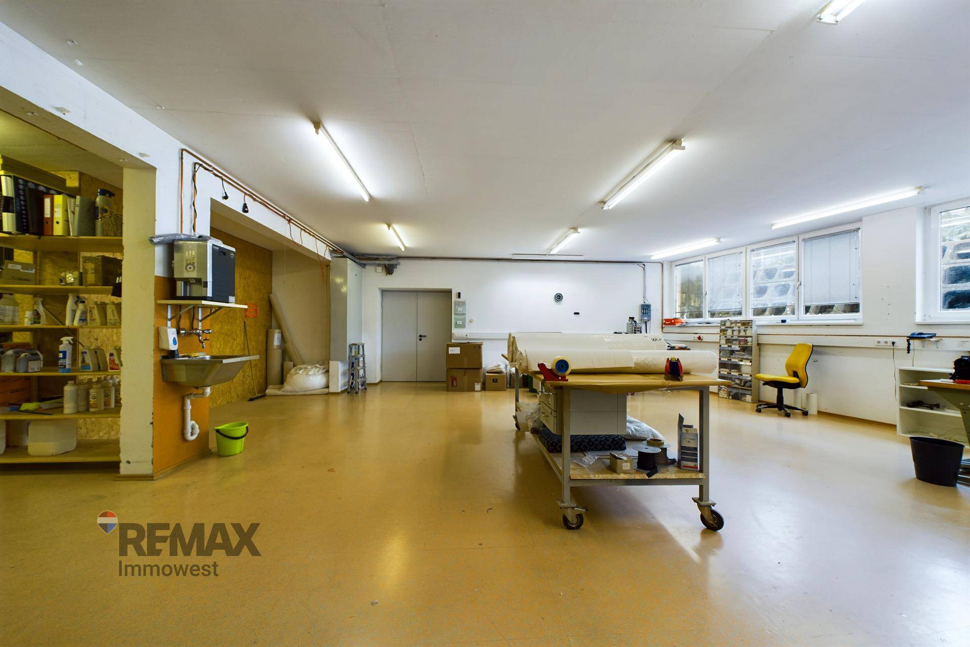Großer Werkstatt- oder Produktionsbereich mit Spülbecken, Arbeitstischen und Fenstern, heller gelber Boden.