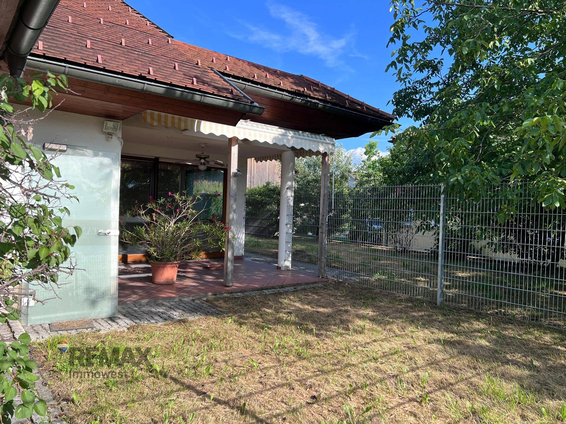 Überdachte Terrasse mit Topfpflanze und angrenzendem, eingezäuntem Gartenbereich.