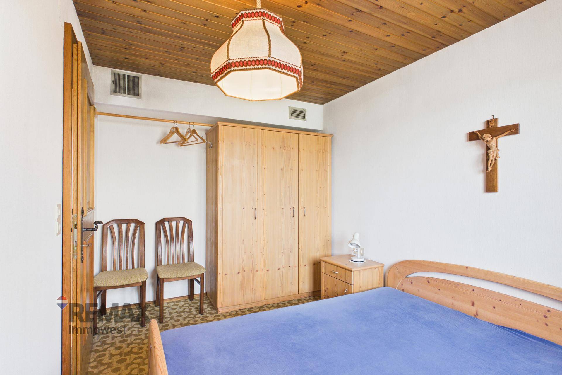 Schlafzimmer mit Einzelbett, Holzschrank und religiöser Dekoration an der Wand.
