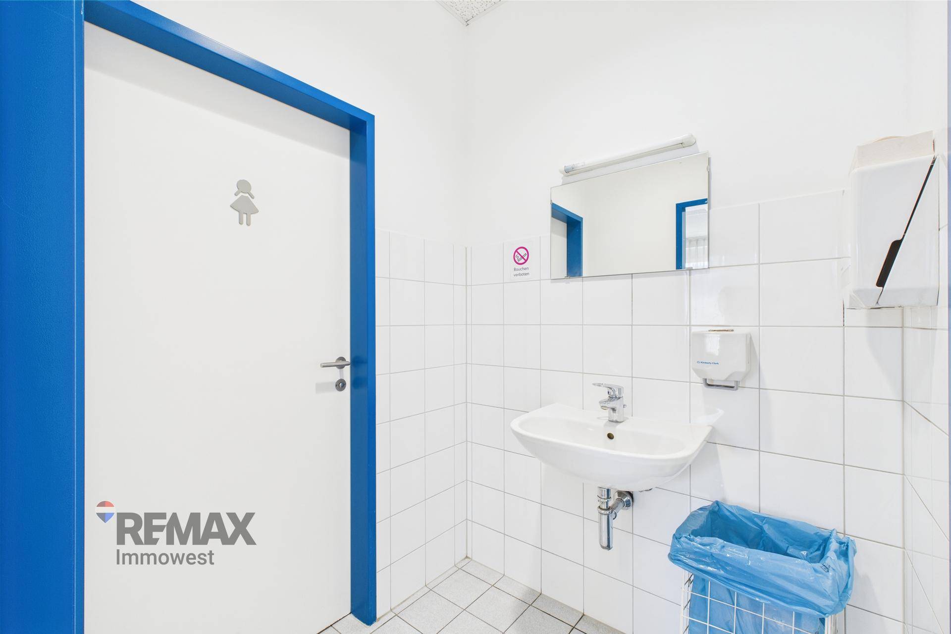 Sauberer Toilettenraum mit weißen Fliesen, Waschbecken und einer Tür mit blauer Zarge.