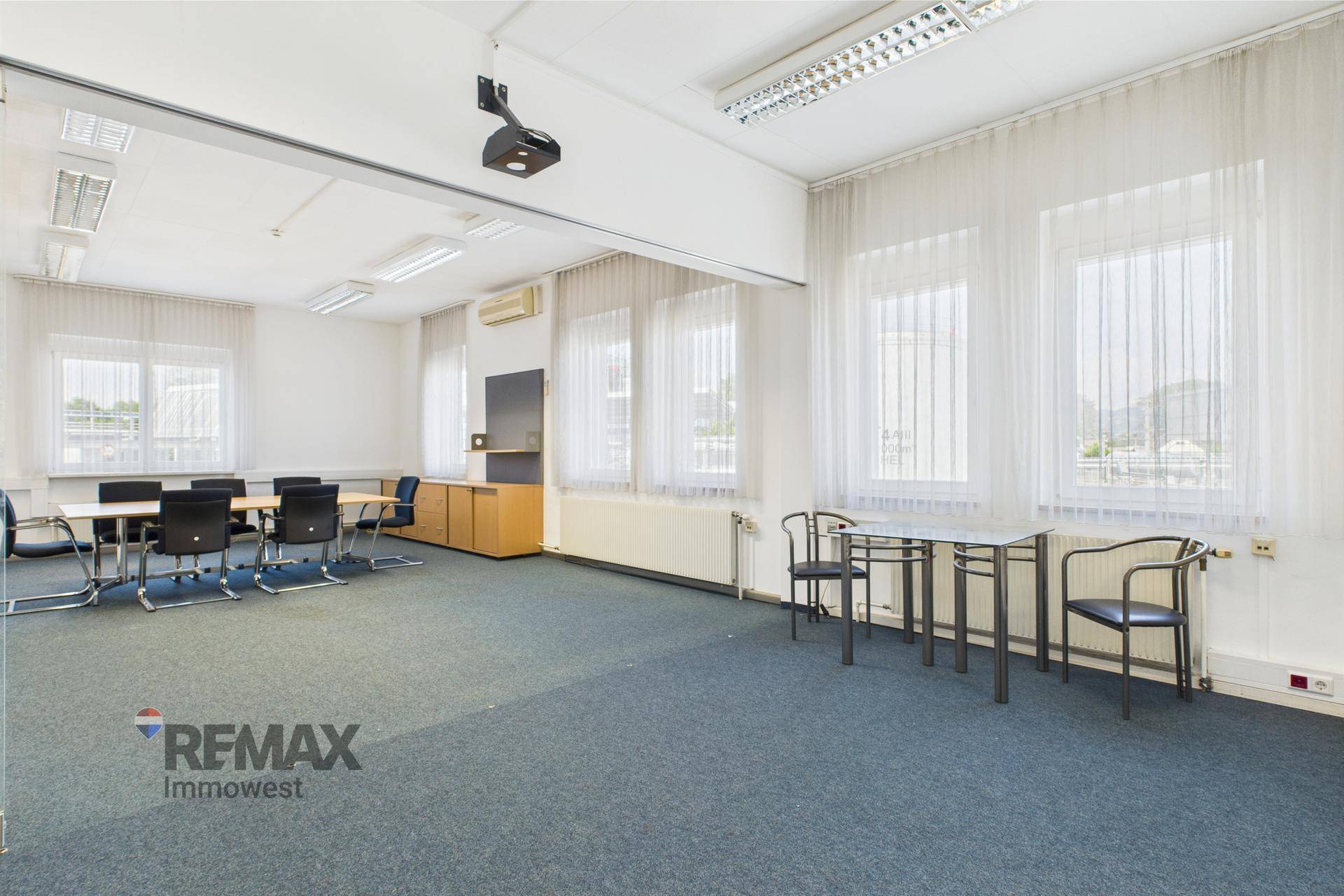 Großer Konferenzraum mit Teppichboden, Besprechungstisch, Stühlen und einem Beamer an der Decke.