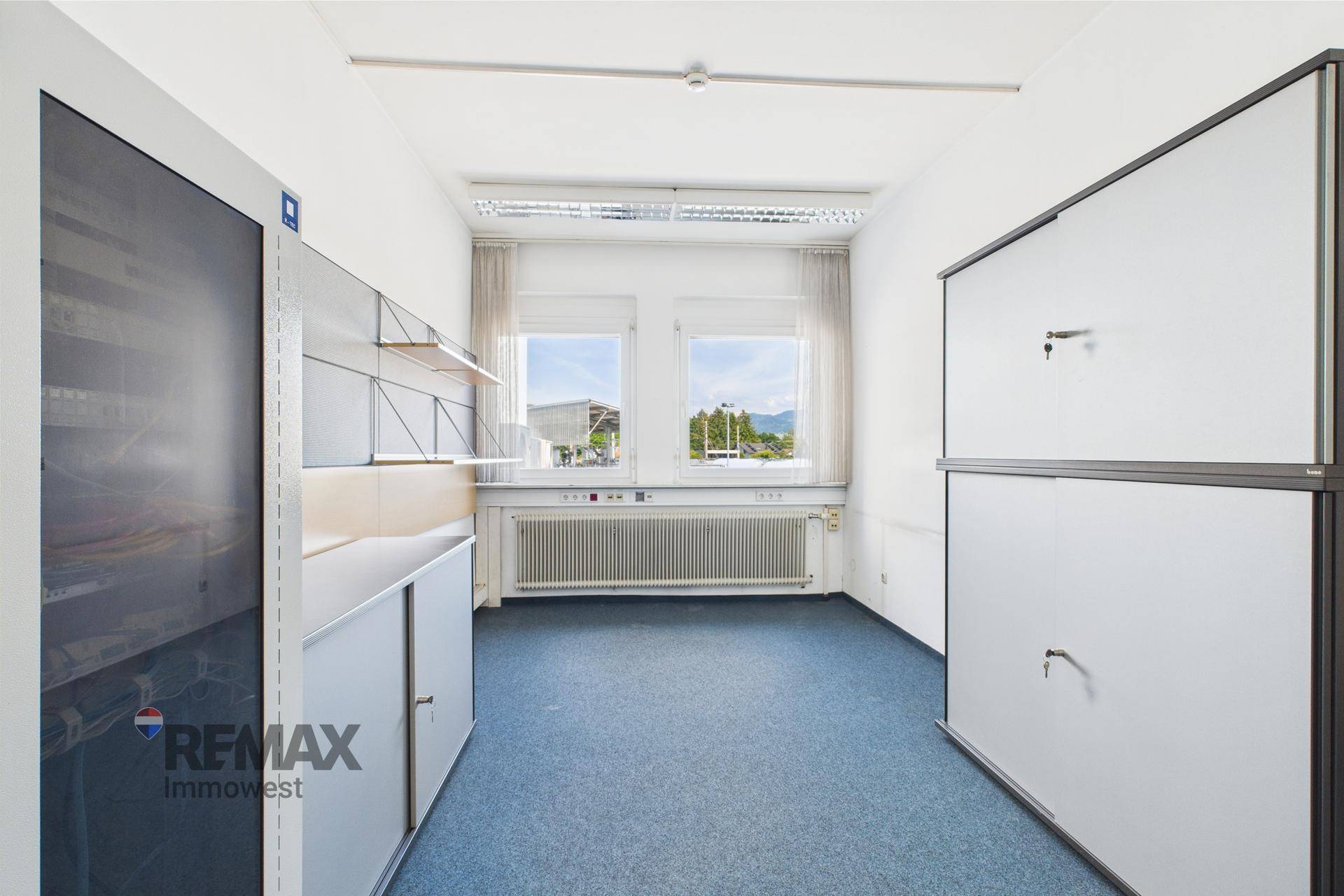 Helles Einzelbüro mit Teppichboden, zwei Fenstern und integrierten Schränken für Stauraum.