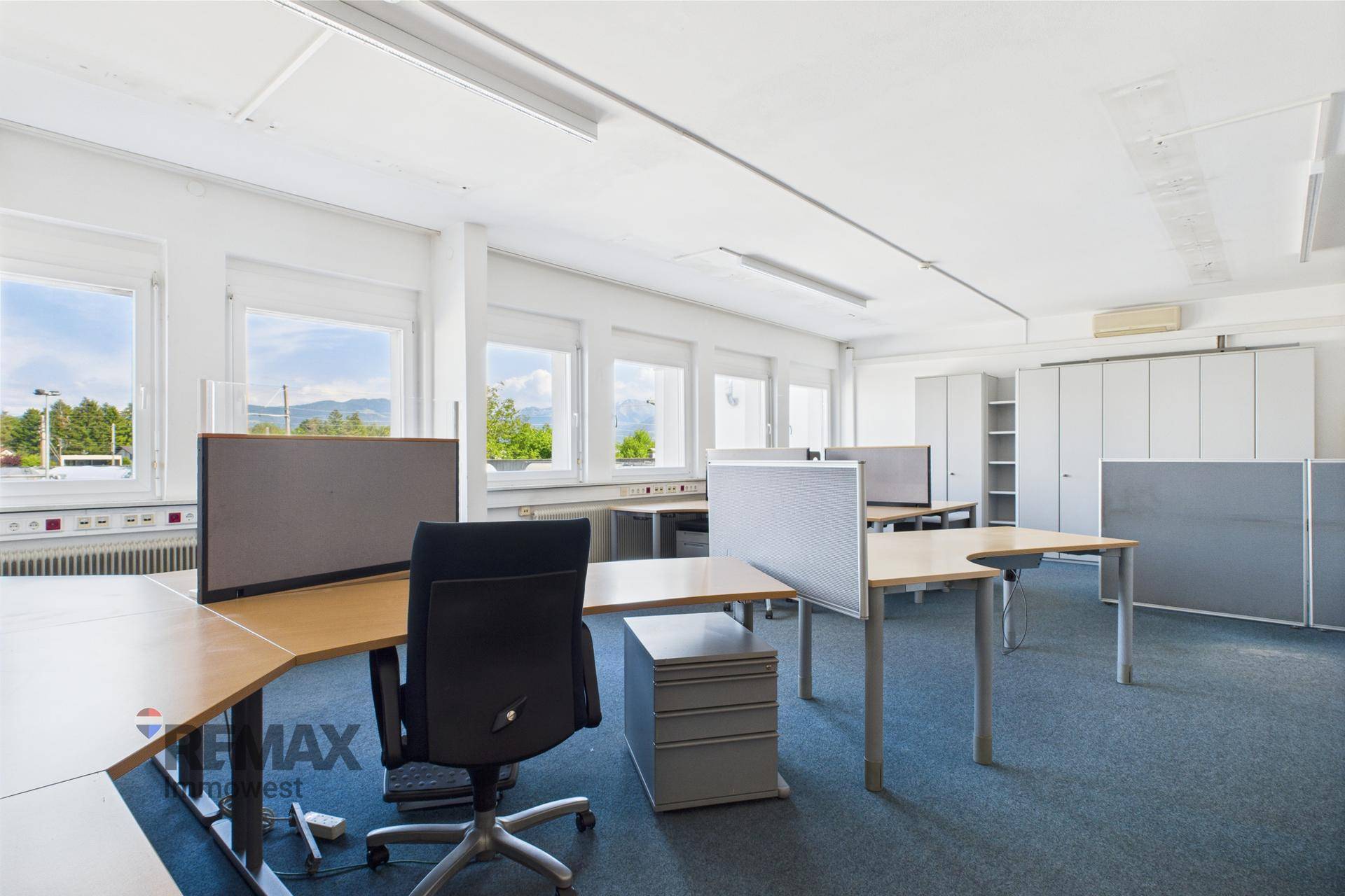 Großraumbüro mit mehreren Arbeitsplätzen, Bürostühlen und Fenstern mit Blick ins Grüne.
